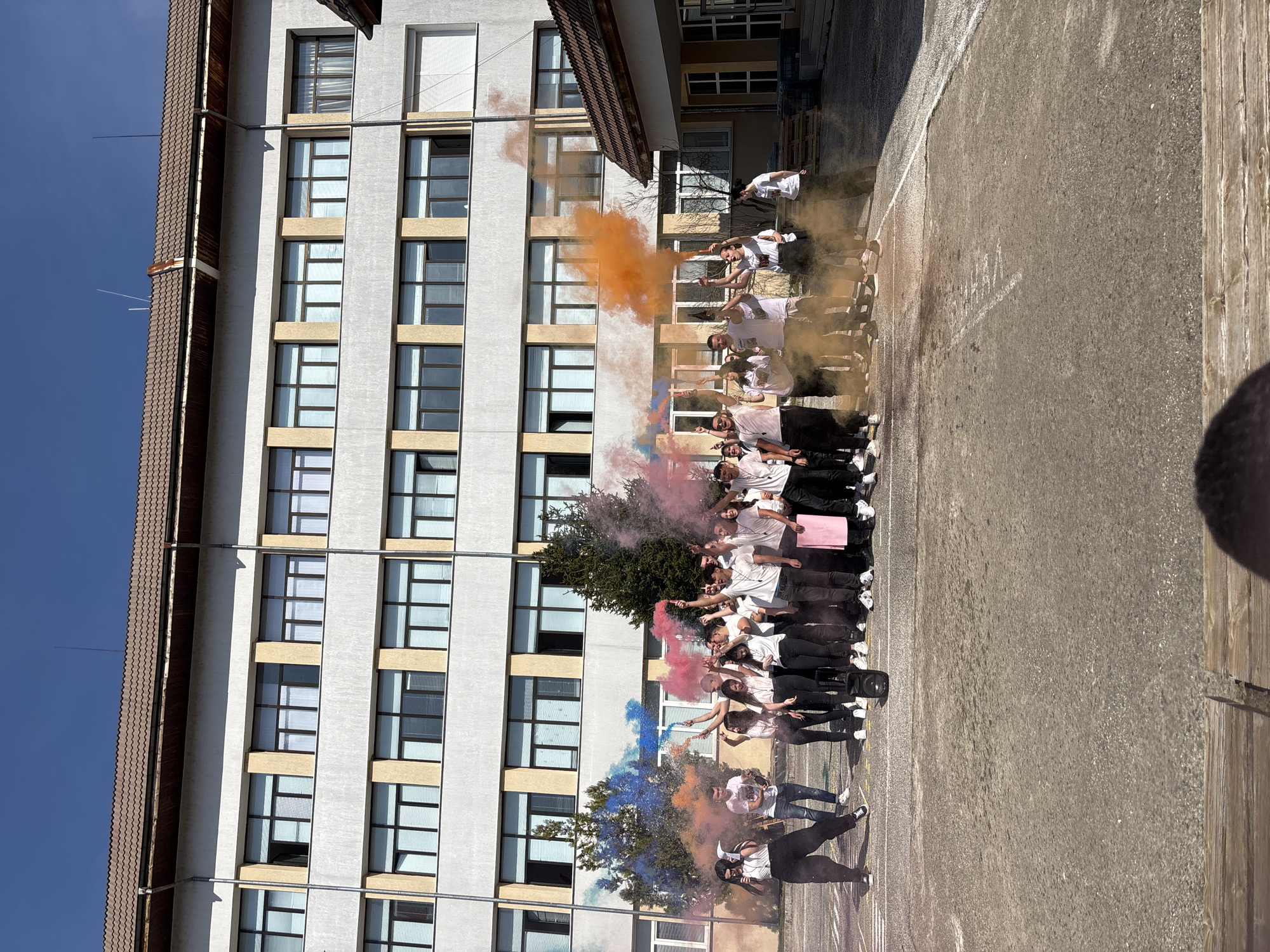 The scene depicts a group of approximately 20 to 25 young individuals gathered in an outdoor paved area, identified as being in the city of Zlatograd, Bulgaria. The background features a multi-story building, likely a school, characterized by a light-colored, textured facade, multiple rectangular windows with dark frames and horizontal blinds, and a brown-tiled roof with visible gutters. The sky above is clear and blue.
The individuals, predominantly wearing white tops and dark bottoms, are distributed across the central and right portions of the frame. Many are actively holding and activating colored smoke emitters, generating plumes of pink, orange, blue, and purple smoke that emanate upwards and outwards around the group. Several individuals have their arms raised in celebratory gestures.
On the far left, one person in a white top and black pants is holding a white rectangular object and appears to be speaking into a handheld microphone. A black portable speaker rests on the ground near the center of the group. Another individual slightly right of center holds a pink rectangular object. Green foliage, possibly trees or large shrubs, is visible behind the group, partially obscured by the smoke. The ground is a light gray asphalt or concrete surface with faint white markings. The scene is illuminated by bright sunlight, casting visible shadows.
