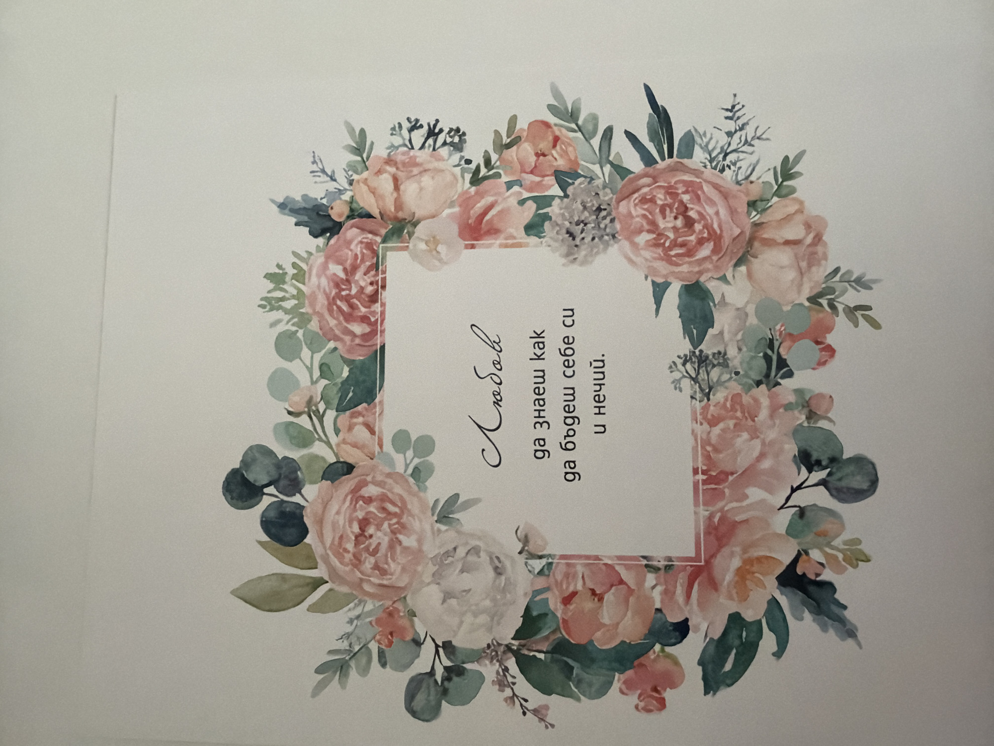 The image is a close-up, flat lay photograph of a printed card with a floral wreath design. The wreath features a watercolor-style depiction of pink and white peonies, eucalyptus leaves, and smaller sprigs of greenery. Inside the wreath, there is a square white space with text in Bulgarian. The text reads "Любов" (Lyubov - Love) at the top, followed by "да знаеш как" (da znaesh kak - to know how), "да бъдеш себе си" (da budesh sebe si - to be yourself), and "и нечий." (i nechiy. - and someone's.). The background of the card is white, and the overall image is well-lit with no visible shadows. The scene depicted is an artistic composition and not a location in the city of Pazardzhik, Bulgaria.