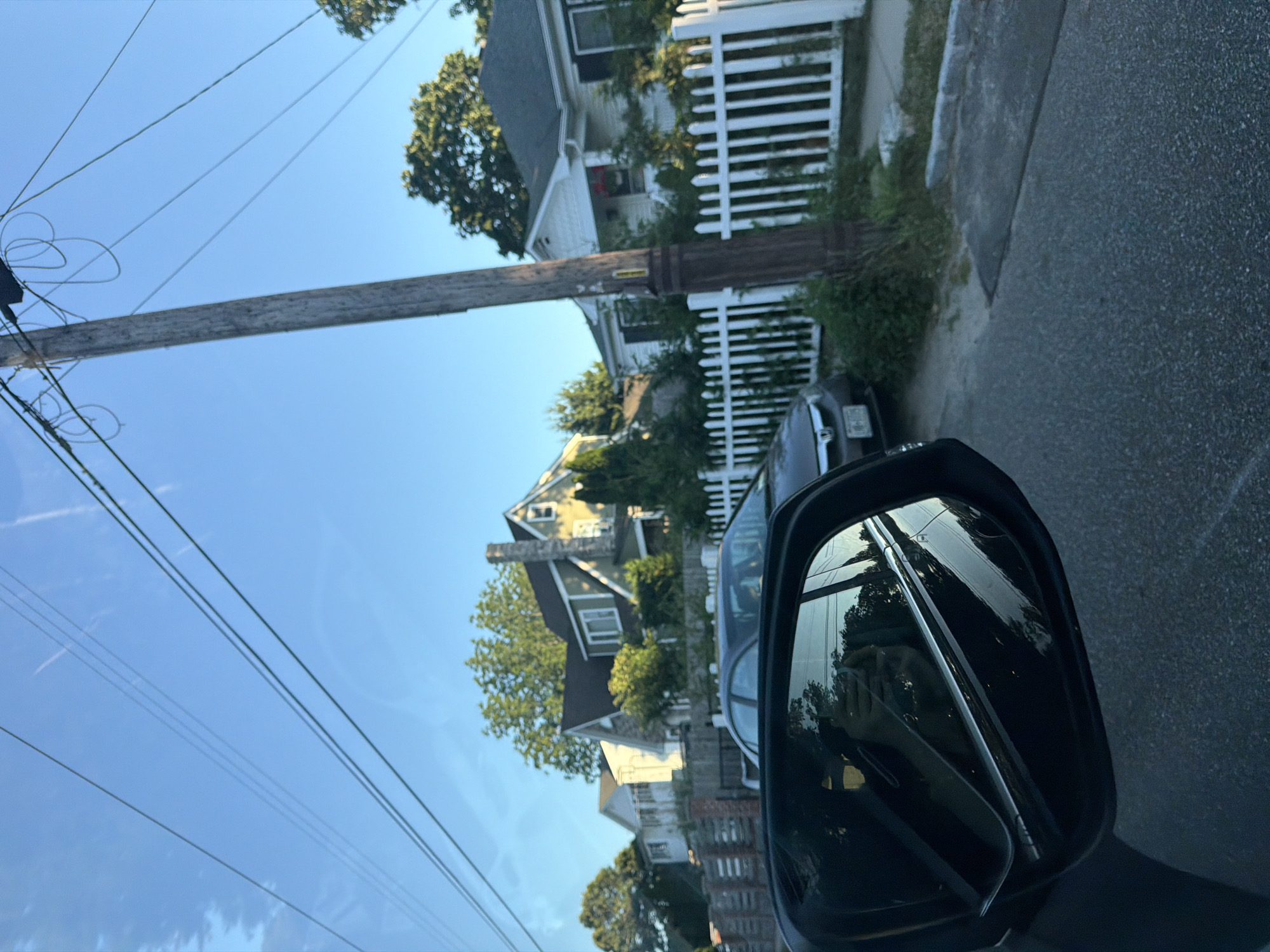 A side-view mirror of a vehicle reflects a person holding a cell phone, capturing the image. The reflection also shows overhead power lines and trees. Outside the mirror, a dark-colored sedan is parked on the right side of the street. The background reveals a residential street in Riverhead, USA, with several houses, a white picket fence, trees, and a utility pole. The sky is clear and blue.