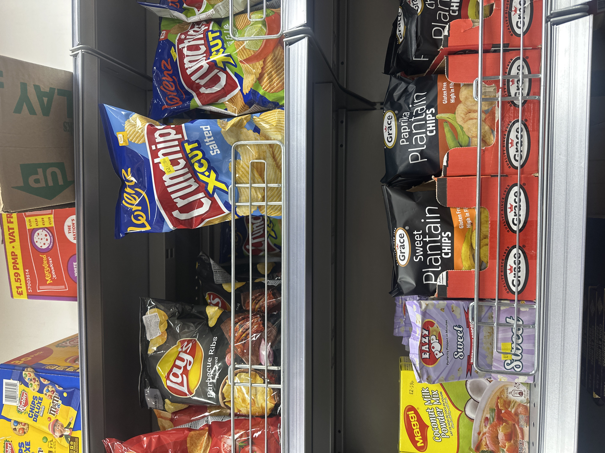 The image shows a section of a metal shelving unit in a retail environment, likely a convenience store or supermarket. The shelves are stocked with various snack foods, predominantly crisps (potato chips).
On the left side of the image, there are bags of Lorenz Crunchips X-Cut, Lay's Barbecue Ribs crisps, Keebler Chips Deluxe Minis and Maryland Chunkies cookies.
On the right side, there are boxes of Grace Plantain Chips in Paprika and Sweet flavors, Eazy Pop Sweet microwave popcorn, and Maggi Coconut Milk Powder Mix.
The shelves are arranged in a way that allows customers to easily see and access the products. The lighting appears to be typical of a retail environment. The photograph lacks contextual clues to determine the location beyond the presence of British brands and pricing in pounds, indicating it is in the United Kingdom.