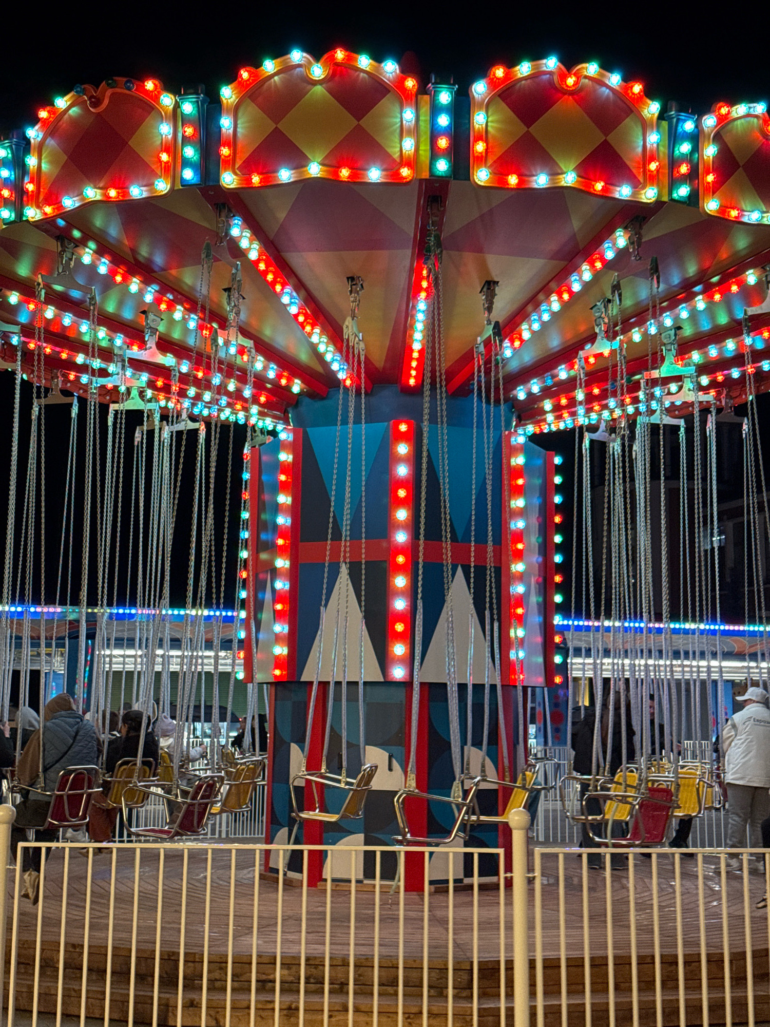 This is a nighttime, low-angle, eye-level photograph of a carousel ride at an amusement park, likely in Tver, Russia. The carousel is adorned with numerous colorful lights, predominantly red, blue, and green, illuminating the structure and its surrounding area. The ride features multiple swings suspended by chains, with some swings occupied by people. The people are indistinct due to the low lighting and distance but appear to be dressed in casual winter attire. The carousel itself is colorful, with a patterned roof and a central pole decorated with geometric designs and lights. A white wooden fence is in the foreground, separating the viewer from the ride. The background is dark, indicating it is nighttime.