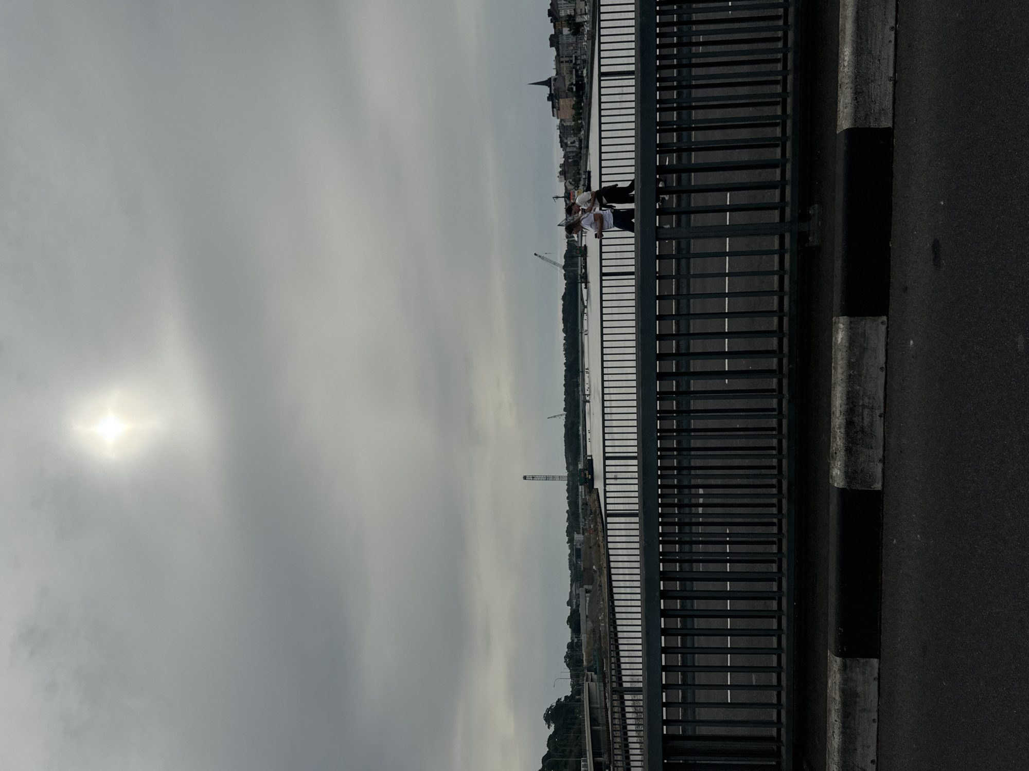 This image depicts a scene from a bridge overlooking the River Suir in Waterford, Ireland. The sky is overcast with the sun shining through the clouds, creating a bright spot. On the bridge's walkway, two individuals are walking side-by-side, with one holding a phone. In the background, across the river, there is a city skyline with buildings and a prominent church spire. Industrial elements such as cranes and construction sites are also visible along the waterfront, suggesting ongoing development. The bridge itself has a metal railing and a road surface marked with white lines. The foreground shows the curb and asphalt of the road on the bridge.