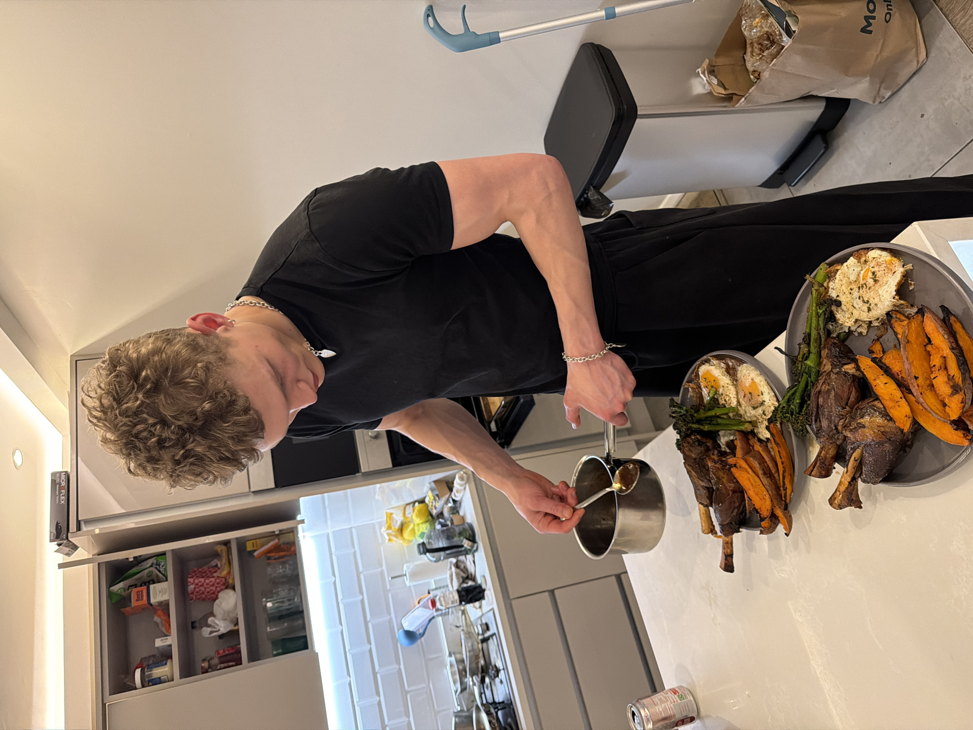 A young male in a black t-shirt and black sweatpants stands in a modern kitchen, pouring sauce from a small saucepan with a spoon onto two plates of food. The plates contain roasted lamb shanks, sweet potato wedges, asparagus, and fried eggs. The kitchen has white cabinets and countertops, with a tiled backsplash. A bin with a bag of groceries sits behind him. The location is likely a residential kitchen, based on the design and contents, but there's no indication it is in the city of London.