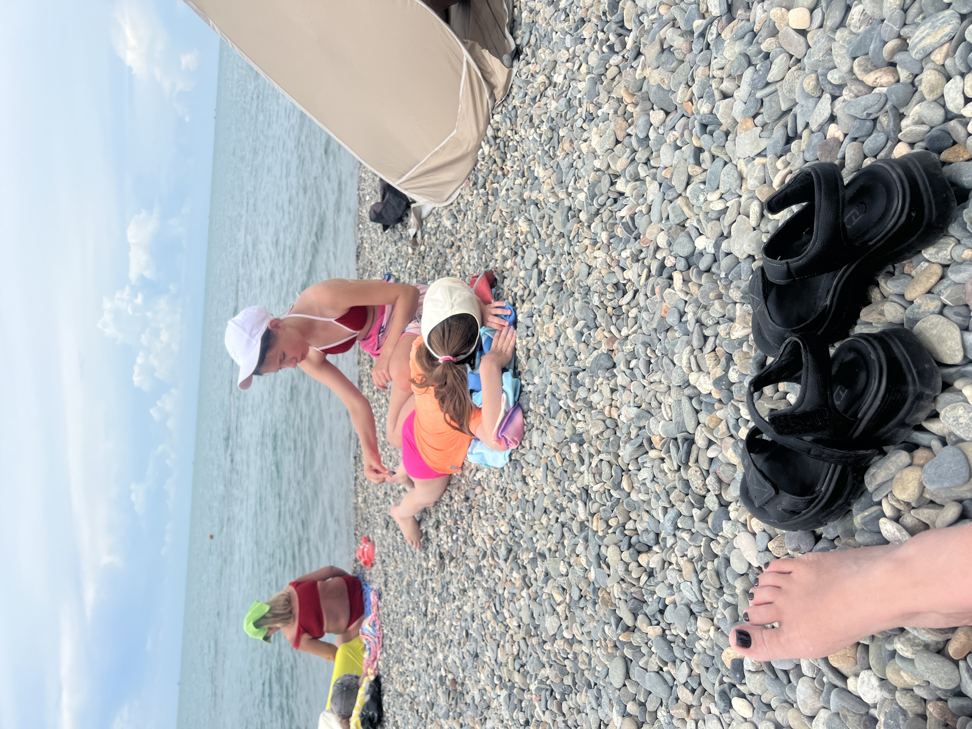 A high-angle medium shot captures a beach scene with four individuals in various states of relaxation on a pebble-covered shoreline. In the foreground, a person's left foot with black nail polish is visible, along with a pair of black sandals. Further into the scene, a young person wearing a white cap and a red bikini top is seated, reaching towards another young person lying on their stomach. This second individual is wearing an orange t-shirt and pink shorts, with their face obscured by a white cap and their body resting on a light blue towel. In the background, a person in a red bikini and a green headscarf sits facing away from the camera, next to a yellow inflatable object. To the right of the frame, a section of a tan beach tent is visible. The sea extends into the distance under a cloudy sky.