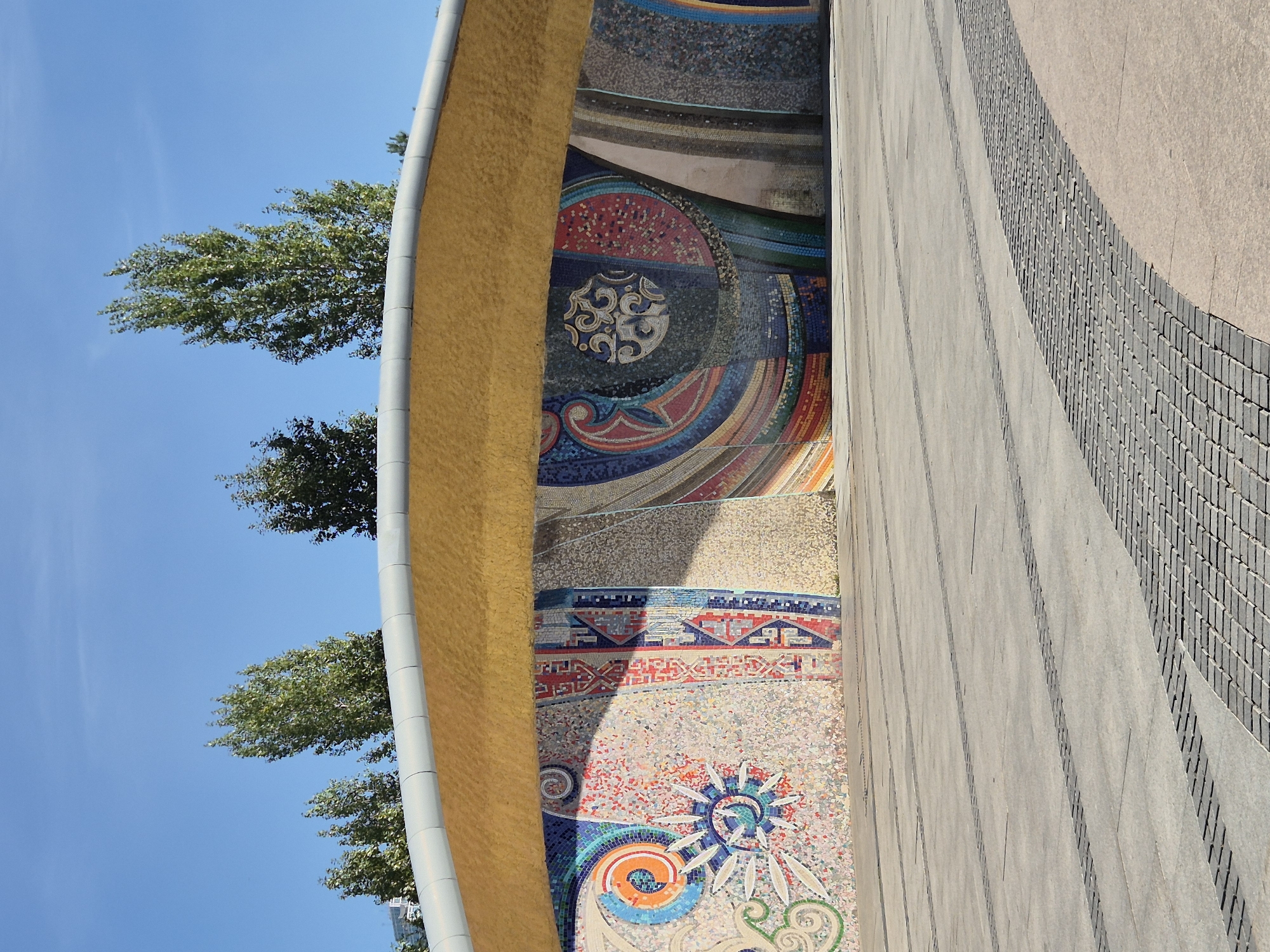 The image shows a wide outdoor plaza in Astana, Kazakhstan, in front of a building with a distinctive curved, gold-colored canopy. Beneath the canopy is a large, colorful mosaic mural that spans the length of the building's facade. The mural features abstract geometric patterns, swirling designs, and sun-like motifs in a variety of vibrant colors including blues, oranges, reds, and yellows. The ground of the plaza is paved with large, light-colored rectangular tiles, interspersed with decorative rows of smaller, dark-colored cobblestones. Several rows of these cobblestones form curved lines across the plaza, creating a visual pattern. The sky is a clear, bright blue, with a few wispy white clouds. Tall, leafy green trees are visible above the canopy of the building. No people are visible in the image. The scene appears to be a public space, likely designed for pedestrian use and visual appreciation of the artwork and architecture.