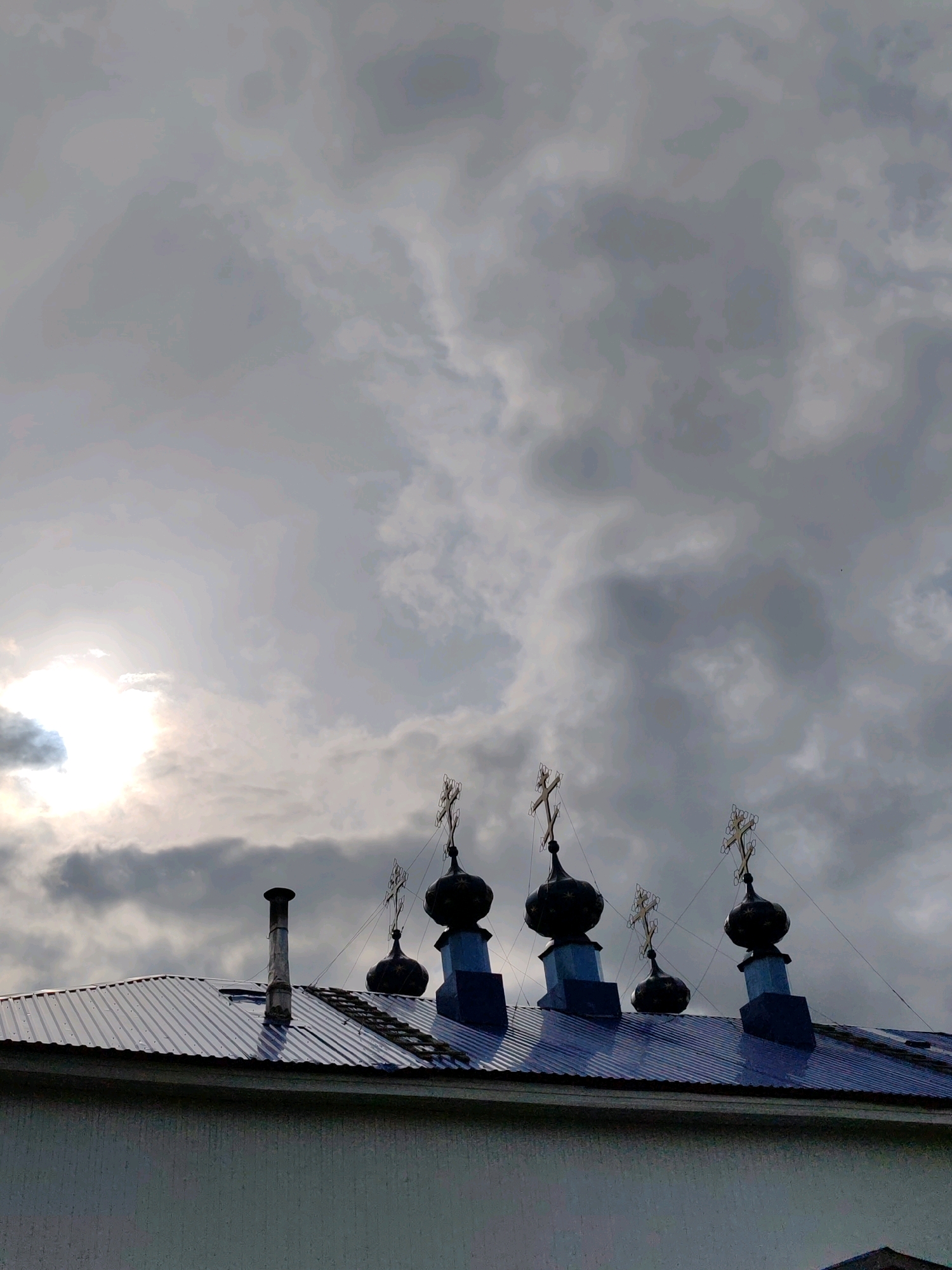 The image captures a portion of a building's rooftop in Chuclen, Russia, under a dramatic, overcast sky. The rooftop is constructed of corrugated metal, reflecting the dim light. Dominating the top of the building are several dark, onion-shaped domes, each topped with a cross. To the left of the domes, a metallic chimney protrudes from the roof. The sun is visible through the heavy clouds on the left side of the frame, casting a bright, diffused glow. The building's facade, visible at the bottom, appears to be a light color with a textured surface. There are no people or animals visible in the frame.
