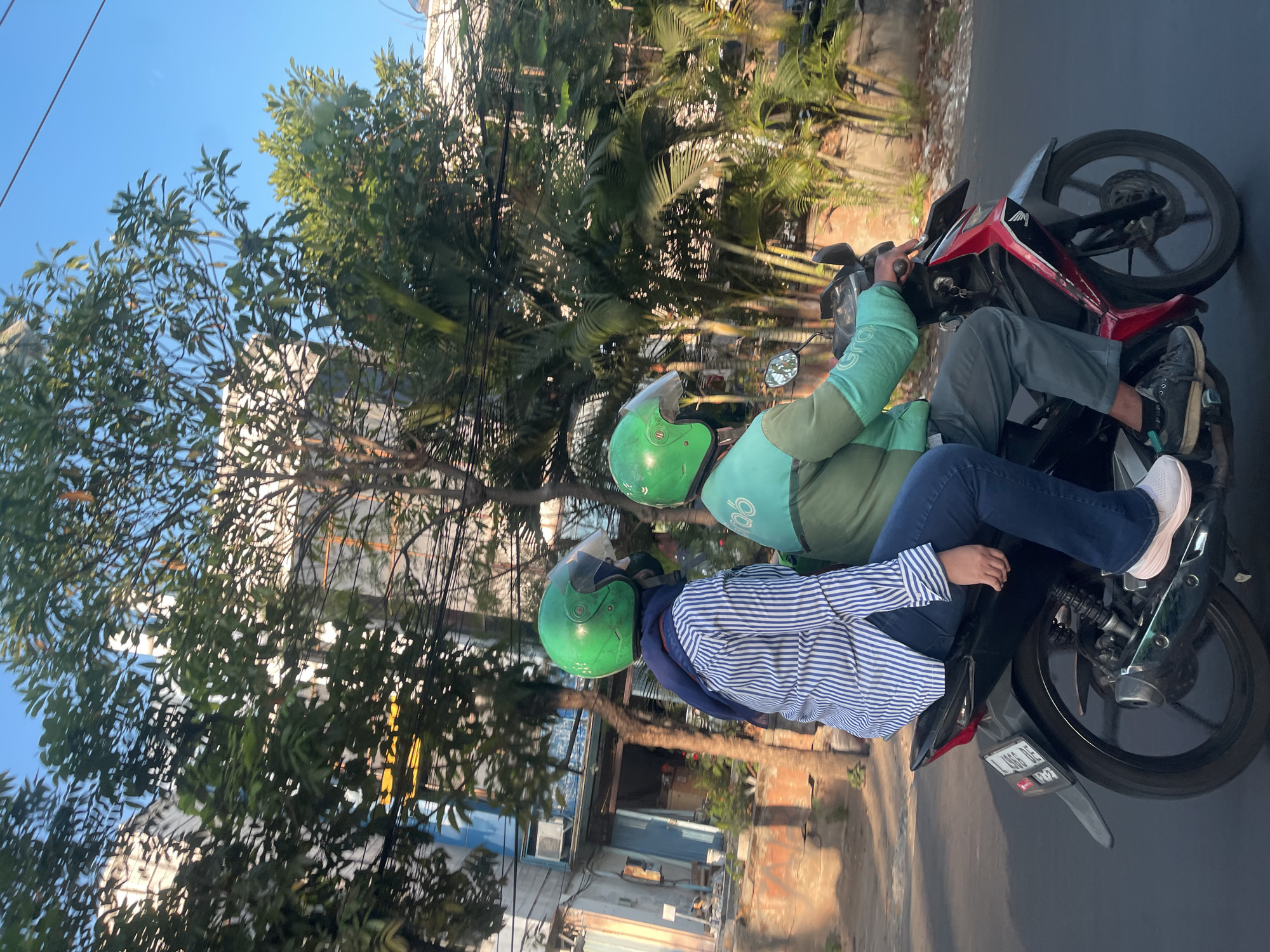 In Surabaya, Indonesia, a motorcycle carrying two individuals rides along a street. The passenger, a woman wearing a blue and white striped shirt, dark blue jeans, and light pink sneakers, is seated behind the driver. She has a dark blue scarf wrapped around her head and shoulders and is wearing a green helmet. The driver is wearing a green jacket with the "Grab" logo visible on it, gray pants, black shoes, and a green helmet. The motorcycle itself is black, red, and silver. In the background, trees and buildings line the street, with some electrical wires overhead. The scene is illuminated by the warm light of the sun, suggesting it is either morning or late afternoon.