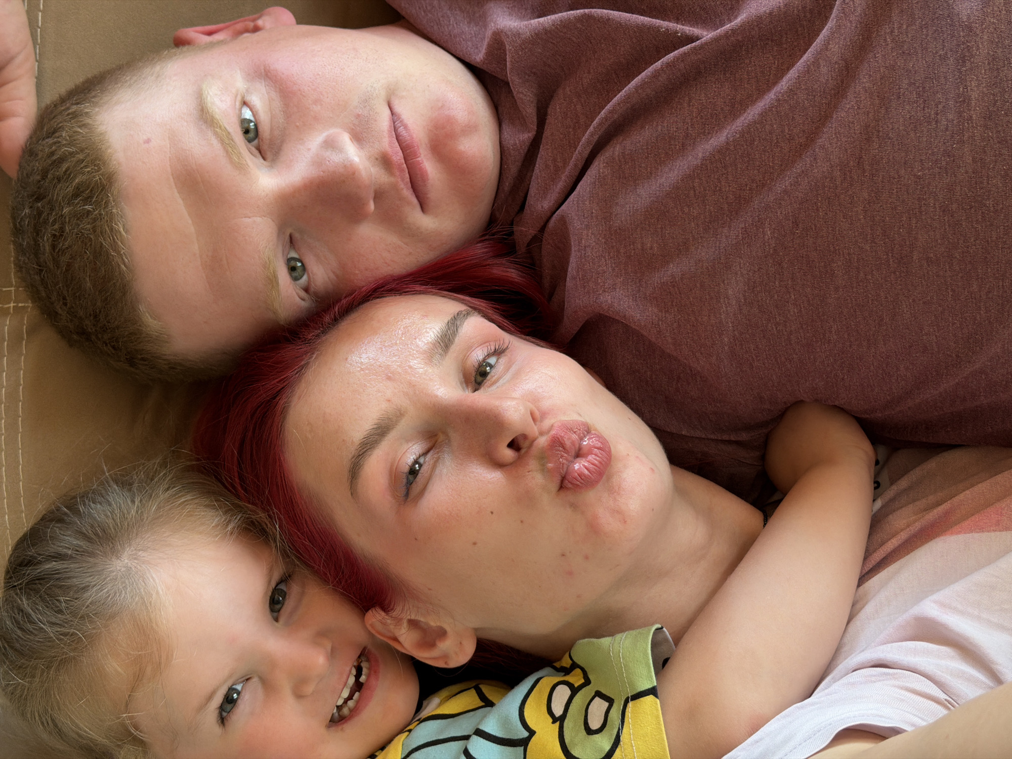 The image is a close-up, overhead shot of three people. On the left is a young child with blonde hair and a bright yellow and blue patterned shirt. The child is smiling with some teeth visible. Next to the child is a woman with bright red hair and a dark pink shirt. She is puckering her lips for a kiss and her eyes are looking towards the camera. To the right of the woman is a man with short brown hair. He is wearing a dark red t-shirt and is looking slightly off-camera with a neutral expression. The people appear to be lying down on a beige surface, possibly a couch or bed. The setting is indoors. There is no visible indication of the city of Chaikovsky, Russia.