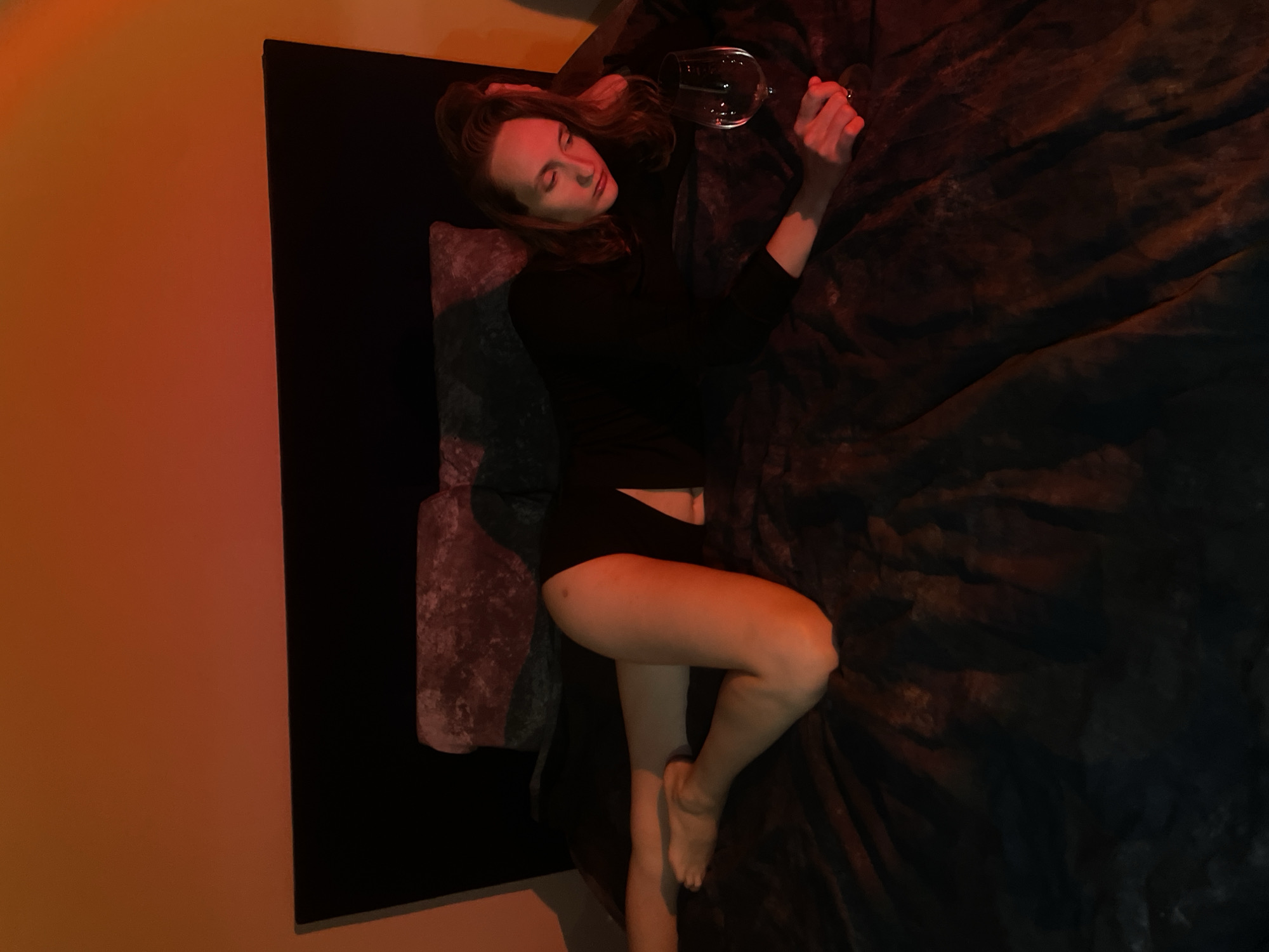 A woman is lying on a textured, dark surface in a room, holding a wine glass. She is wearing a long-sleeved black top and dark shorts. The lighting in the room is dim, with an orange glow on the wall to the left. The photo was taken at Подшивалово, Russia.