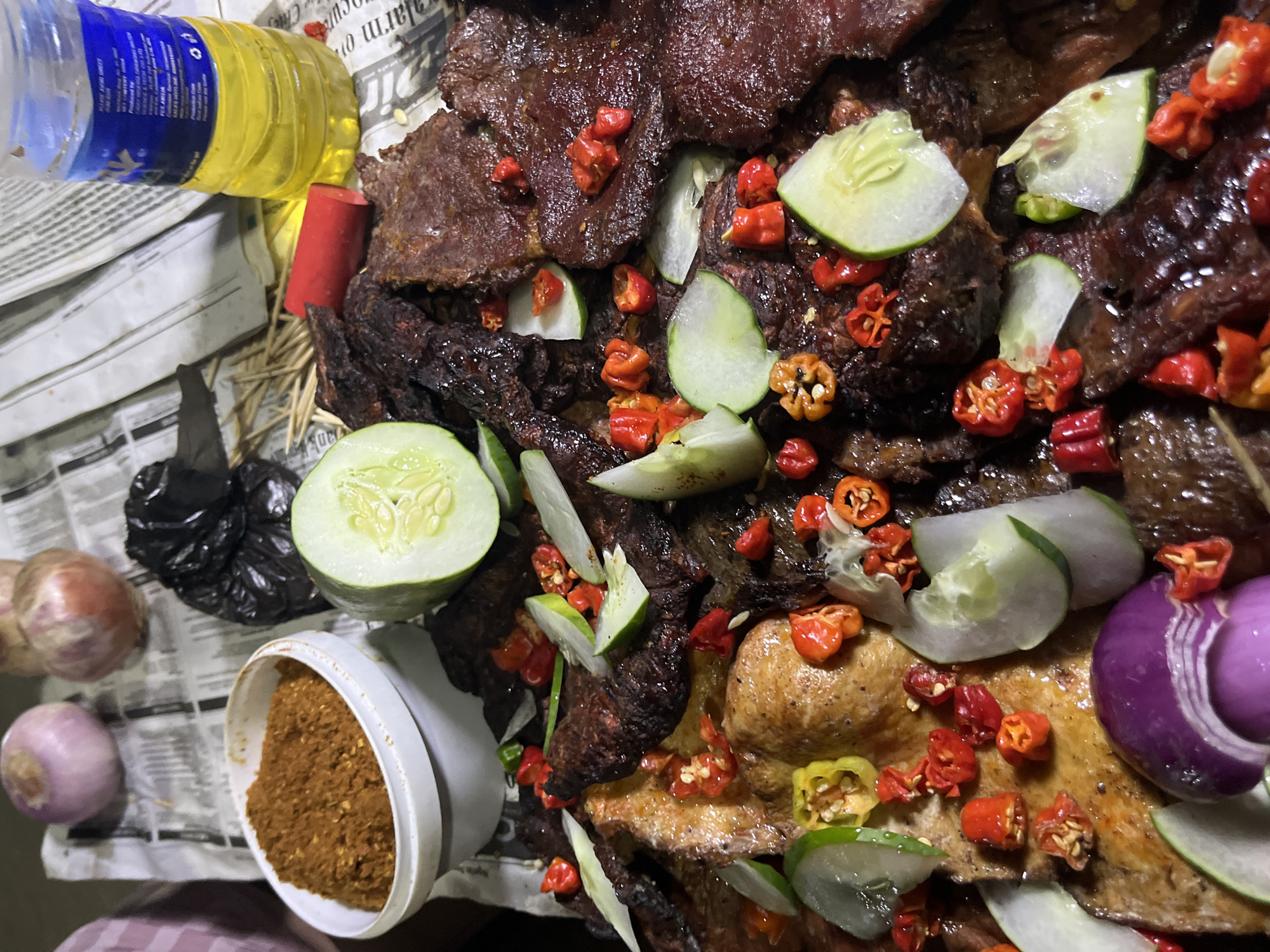 In Abuja, Nigeria, a close-up, overhead shot captures a platter of grilled meat adorned with sliced red chilies and cucumber. The meat, a deep brown hue, appears to be beef jerky or a similar dried and grilled preparation, some pieces thicker than others. Scattered amongst the meat are numerous small, vibrant red bird's eye chilies, some whole and some halved, revealing their seeds. Thin, translucent slices of cucumber are also distributed across the platter, offering a visual contrast to the meat and chilies.
In the background, a white plastic container holds a generous amount of reddish-brown spice mix, possibly a pepper blend or seasoning. Two whole, purple onions sit behind the spice container, one appearing slightly larger than the other. A crumpled black plastic bag is situated to the left of the onions. A small, red cylindrical object, possibly a lighter or a piece of food, lies near a cluster of wooden skewers. To the right, a partially visible plastic bottle filled with yellow liquid, likely oil or a beverage, stands upright. The entire scene is laid out on a surface covered with what appears to be newspaper, adding a rustic texture to the presentation. The lighting is natural and somewhat diffused, highlighting the textures and colors of the food.