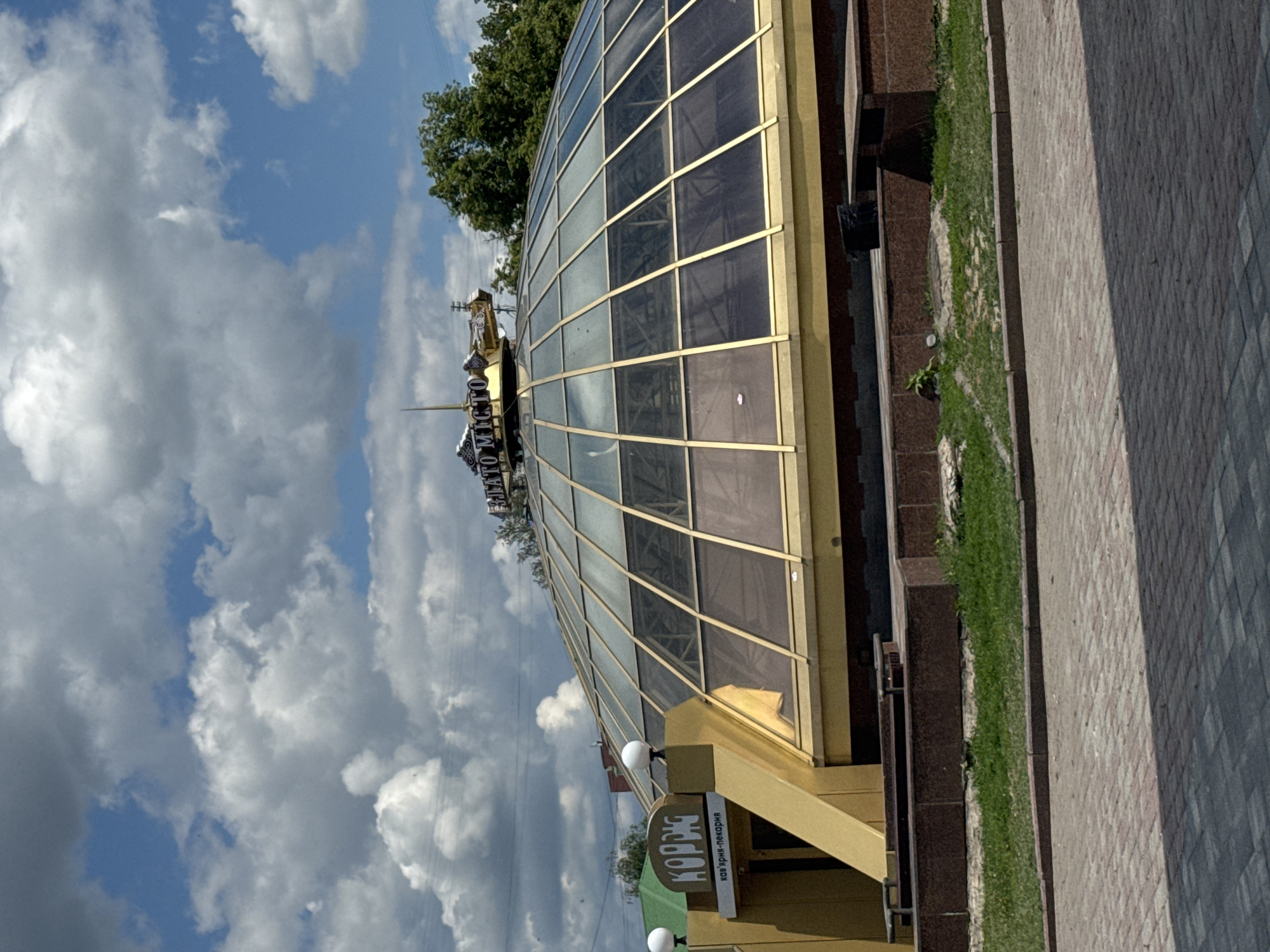 The image captures a daytime scene featuring a modern building with a distinctive architectural style. The most prominent feature is a large, curved glass roof structure, resembling a dome, supported by golden or brass-colored frames. The glass panels appear to be tinted, reflecting the sky. Below this glass section, the building continues with a golden-toned facade.
A rectangular sign is affixed to the building, featuring text in Cyrillic script. The main text reads "КОРБУС" (KORBUS), with smaller text beneath it stating "кафе-пекарня" (cafe-bakery), indicating the establishment's nature. Two spherical lamps are visible on either side of this sign, suggesting it might be illuminated at night.
To the right of the building, there's a paved pedestrian area, likely a sidewalk, constructed from interlocking grey and reddish pavers. Separating the building from the paved path is a narrow strip of well-maintained green grass, bordered by dark, possibly granite, curbs or planters.
In the background, beyond the building, are lush green trees, suggesting nearby vegetation. The sky above is partly cloudy, with patches of clear blue visible between numerous fluffy white and grey clouds. The overall lighting indicates it is daytime, with soft shadows. No people are visible, and no specific activities are taking place in the static scene.