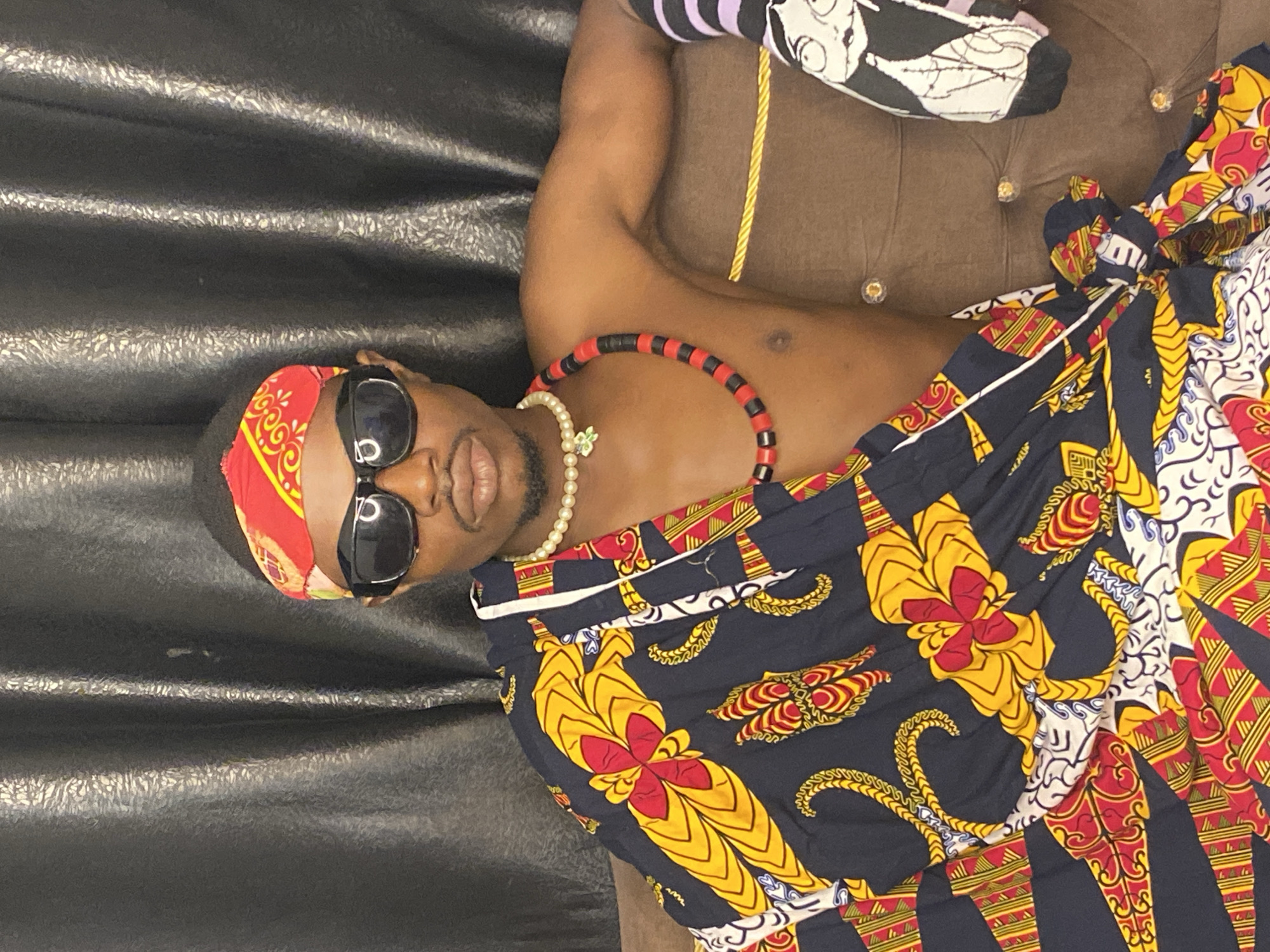 A young Black male with dark skin and a goatee is seated on a brown, tufted sofa against a black, textured backdrop. He is wearing a red and yellow bandana on his head, black sunglasses, and a pearl necklace with a green pendant. Around his neck, he also wears a red and black beaded necklace. A colorful, patterned cloth with yellow, red, and blue designs is draped over his left shoulder and across his chest. His right arm rests on the sofa, and he is wearing a black and purple striped sock on his right arm. The sofa has decorative gold trim and clear button accents. A black doll with white and grey detailing is visible on the right side of the sofa. The scene appears to be indoors, possibly a home setting, in Abakaliki, Nigeria.