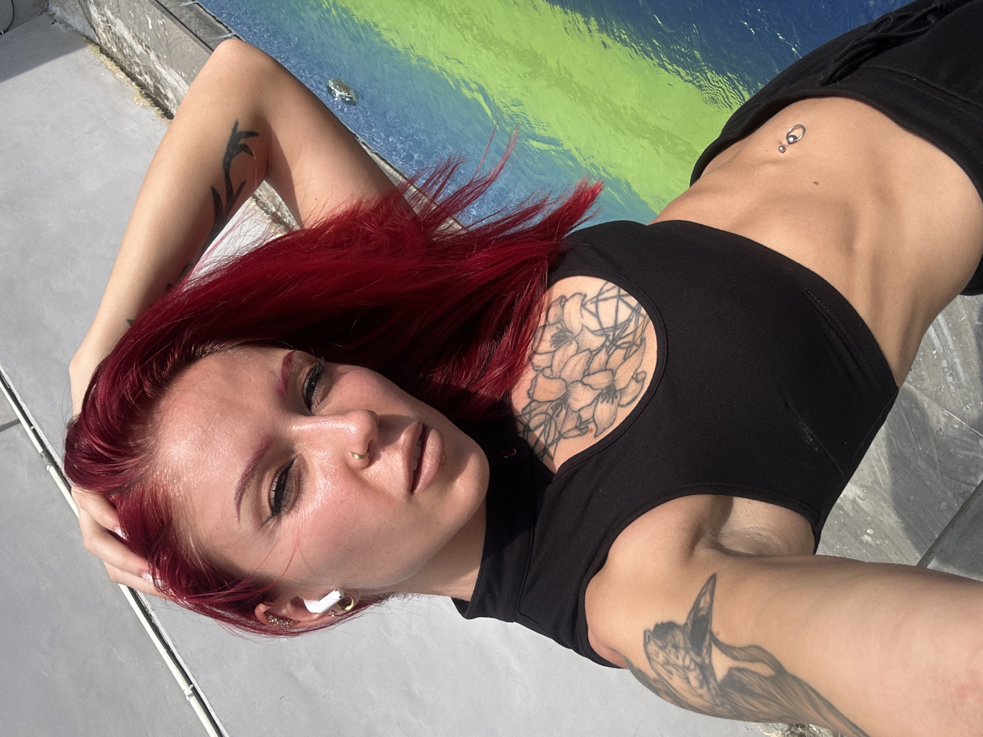 A female individual is captured in an upward-angled selfie, under direct sunlight, in Kerobokan, Indonesia. She has bright red hair and maintains direct eye contact with the camera. Her right hand is placed in her hair, above her head. She wears a black high-necked, sleeveless crop top with a chest cut-out, and black lower garments. Visible tattoos include a floral design on her upper left chest, a large bird on her upper left arm, and a branch pattern on her right forearm. Piercings include a gold nose ring, multiple gold and jeweled left ear piercings, and a navel ring. A white wireless earbud is present in her left ear. The background consists of a light grey wall with a horizontal white conduit on the left, transitioning to a painted surface on the right. This surface displays horizontal bands of bright green above bright blue, exhibiting a semi-reflective texture, with a notable specular highlight on the blue section. The lighting suggests an outdoor environment with strong illumination.