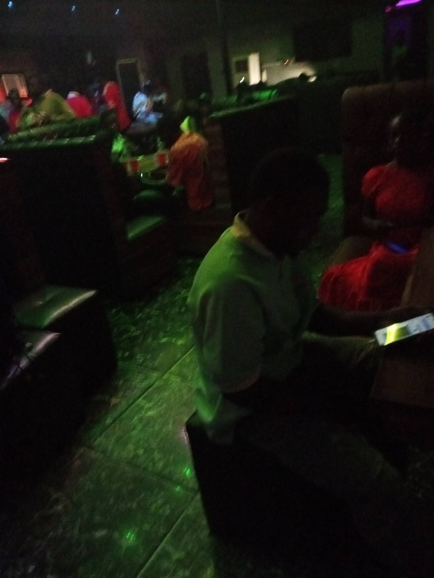 The image appears to have been taken in a dimly lit indoor space, possibly a bar or lounge, located in the city of Unknown, Unknown. Several individuals are present, seated in upholstered chairs or sofas. The floor has a green hue, likely due to ambient lighting. One individual in the foreground is wearing a green top and appears to be looking at a mobile device. Another person, wearing red, is seated beside them. Other patrons are visible in the background.
