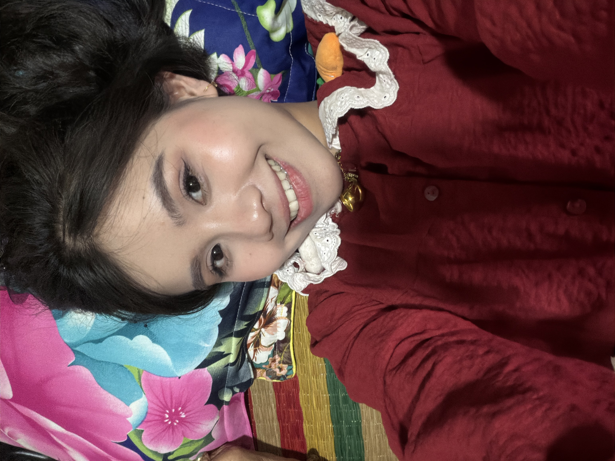 A young woman, likely in Bình An, Vietnam, is captured in a close-up, high-angle selfie, looking directly at the camera with a wide, cheerful smile. She has dark hair, dark eyes, and fair skin, and her head is resting on a patterned surface. She is wearing a dark red long-sleeved top or dress with a prominent white, ruffled collar featuring intricate eyelet lace. A small golden bell charm is visible on her clothing near the collar, and a miniature orange carrot-shaped object is tucked into the right side of her collar.
The background consists of several patterned fabrics. To her upper left is a dark blue fabric with large pink and green floral designs. Below her, to the right, is a colorful striped woven mat, characteristic of Southeast Asian households, displaying vertical bands of red, orange, yellow, green, and blue. A smaller floral pattern with a yellow trim is also partially visible between these two main background elements. The lighting suggests an indoor setting, possibly under artificial light, contributing to a warm overall tone. No text is visible in the image.