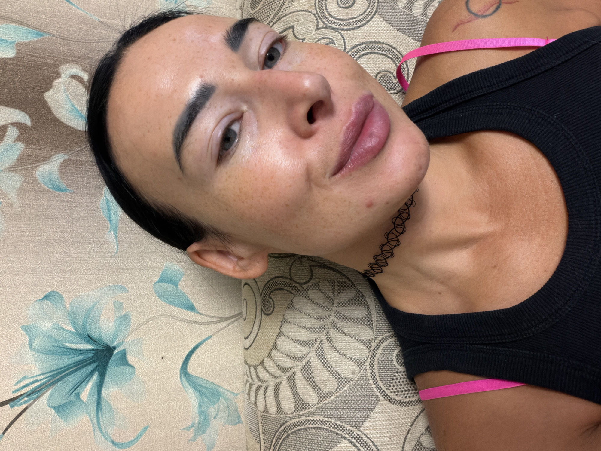 A woman with dark hair is lying on a patterned surface, likely a couch or bed. She is wearing a black tank top with pink straps. She has fair skin with freckles, dark eyebrows, and full lips. She is wearing a black choker necklace. The background consists of a patterned fabric with floral designs. There is no indication of the location being Бронница, Russia.