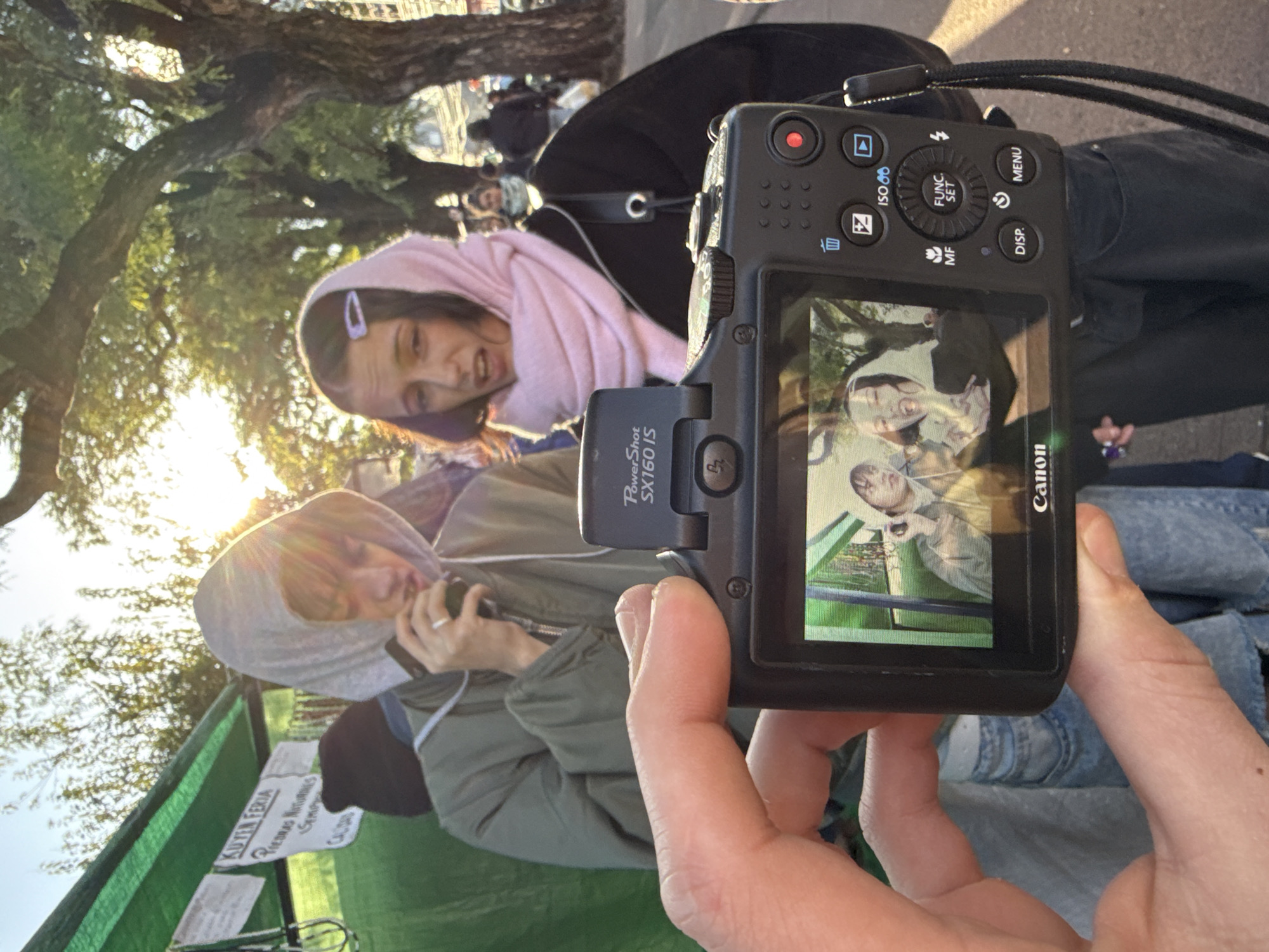 The scene depicts two individuals, one in a pink hoodie and another in a green hooded jacket, standing outdoors, likely at a fair or market stall with a green tent covering it with Spanish text. A hand holds a Canon PowerShot SX160 IS camera displaying a preview of an image captured. The background includes trees, potentially suggesting a park setting within a city. The image is likely taken in Buenos Aires, Argentina, based on the Spanish text on the sign.