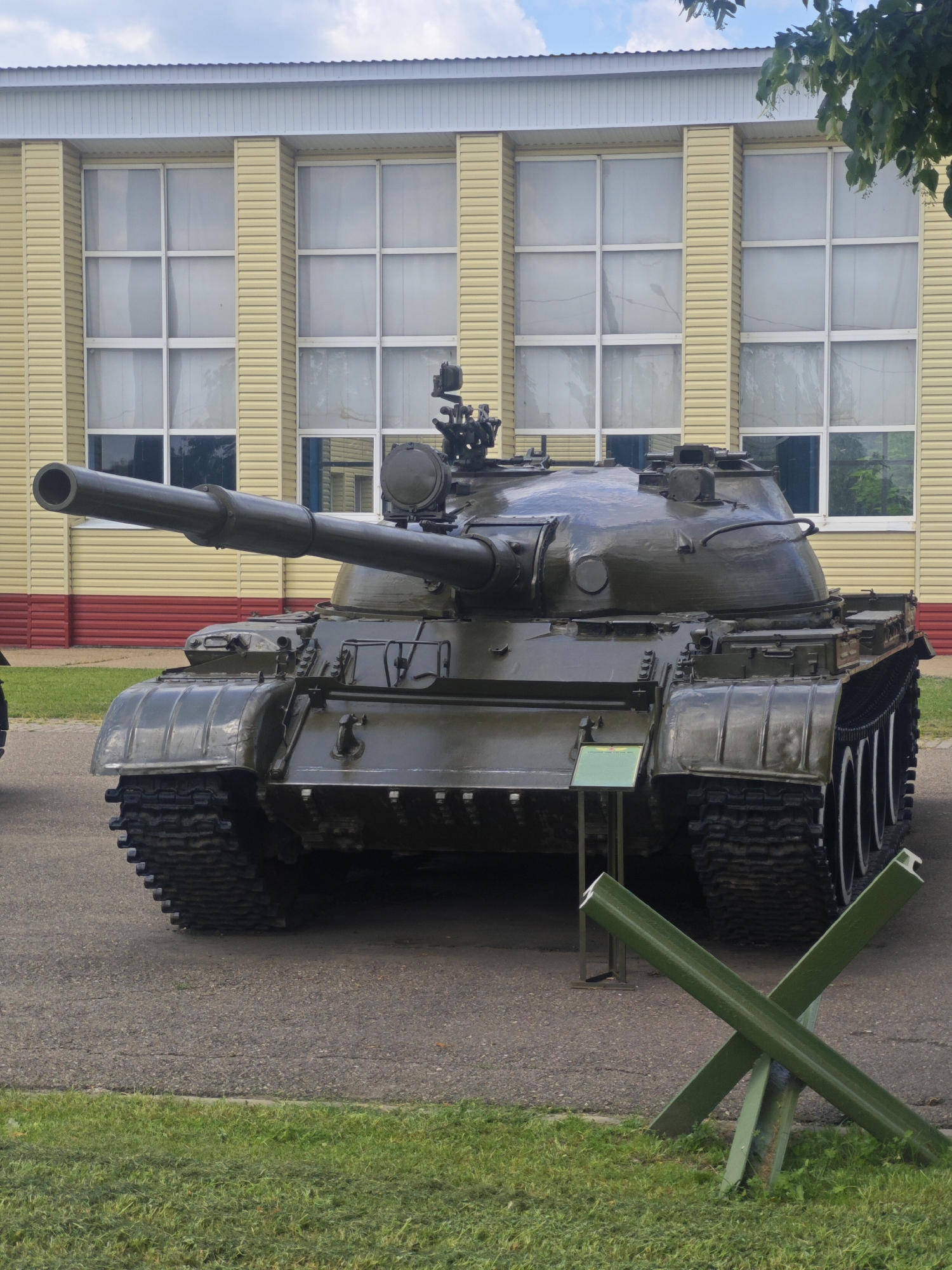 A dark green, tracked military tank is parked outdoors on an asphalt surface in front of a yellow building with multiple large windows. The building has a white roofline and a red band along its lower exterior. In the foreground, to the right of the tank, are two green metal barriers resembling anti-tank obstacles. A patch of green grass is visible in the lower portion of the image. The sky above is partly cloudy. The tank is the primary subject and is positioned centrally, facing the viewer, with its cannon barrel extending to the left. There are no people visible in the frame. The overall scene suggests an outdoor exhibit or display.