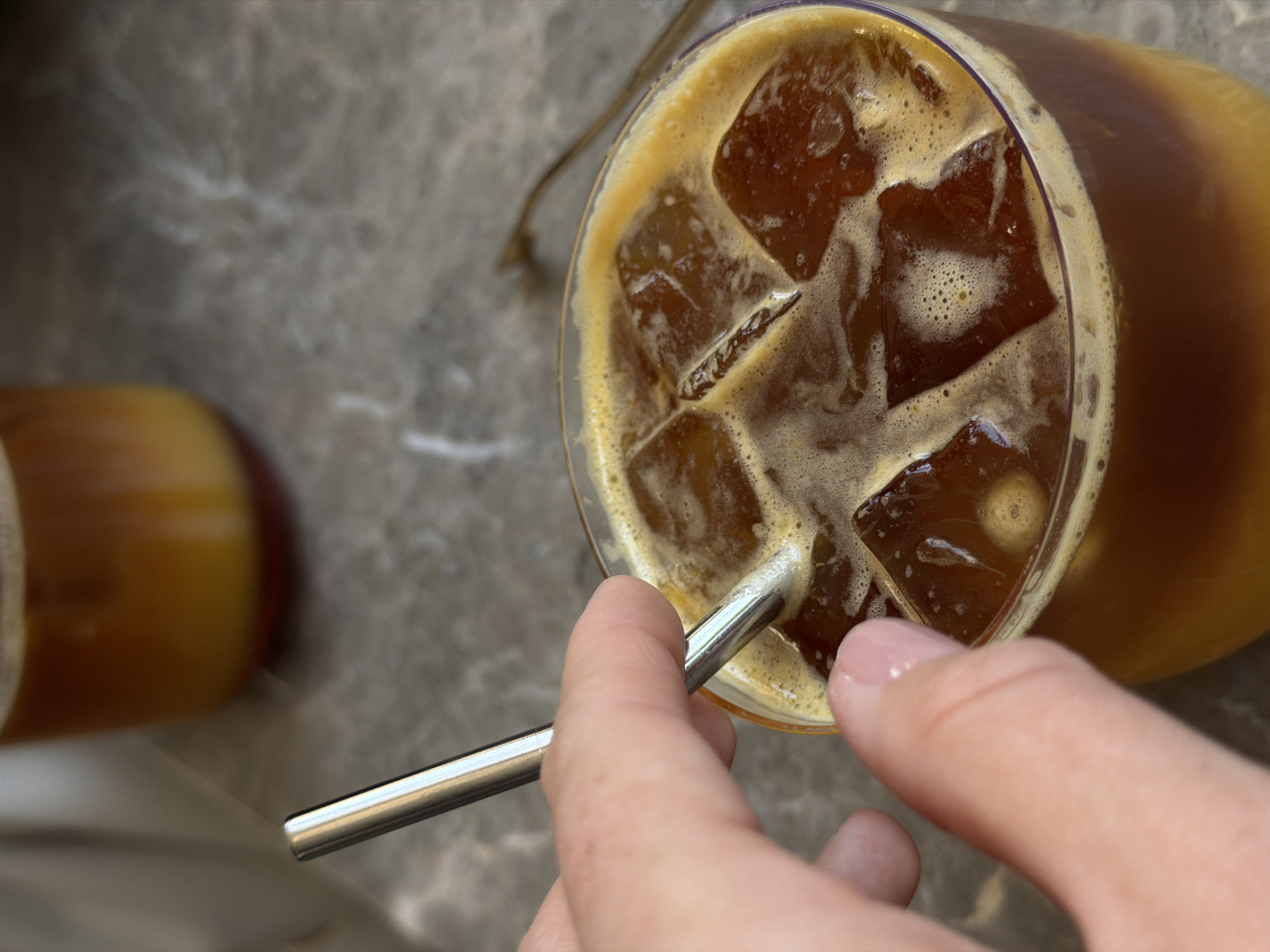 A high-angle shot shows a hand inserting a silver metal straw into a glass of iced coffee on a gray surface. The coffee contains numerous ice cubes and a foamy top. A cylindrical object with a light brown and brown gradient is visible in the blurred background to the left of the glass. The location is plausibly Bishkek City, Kyrgyzstan, but no explicit location cues are visible.