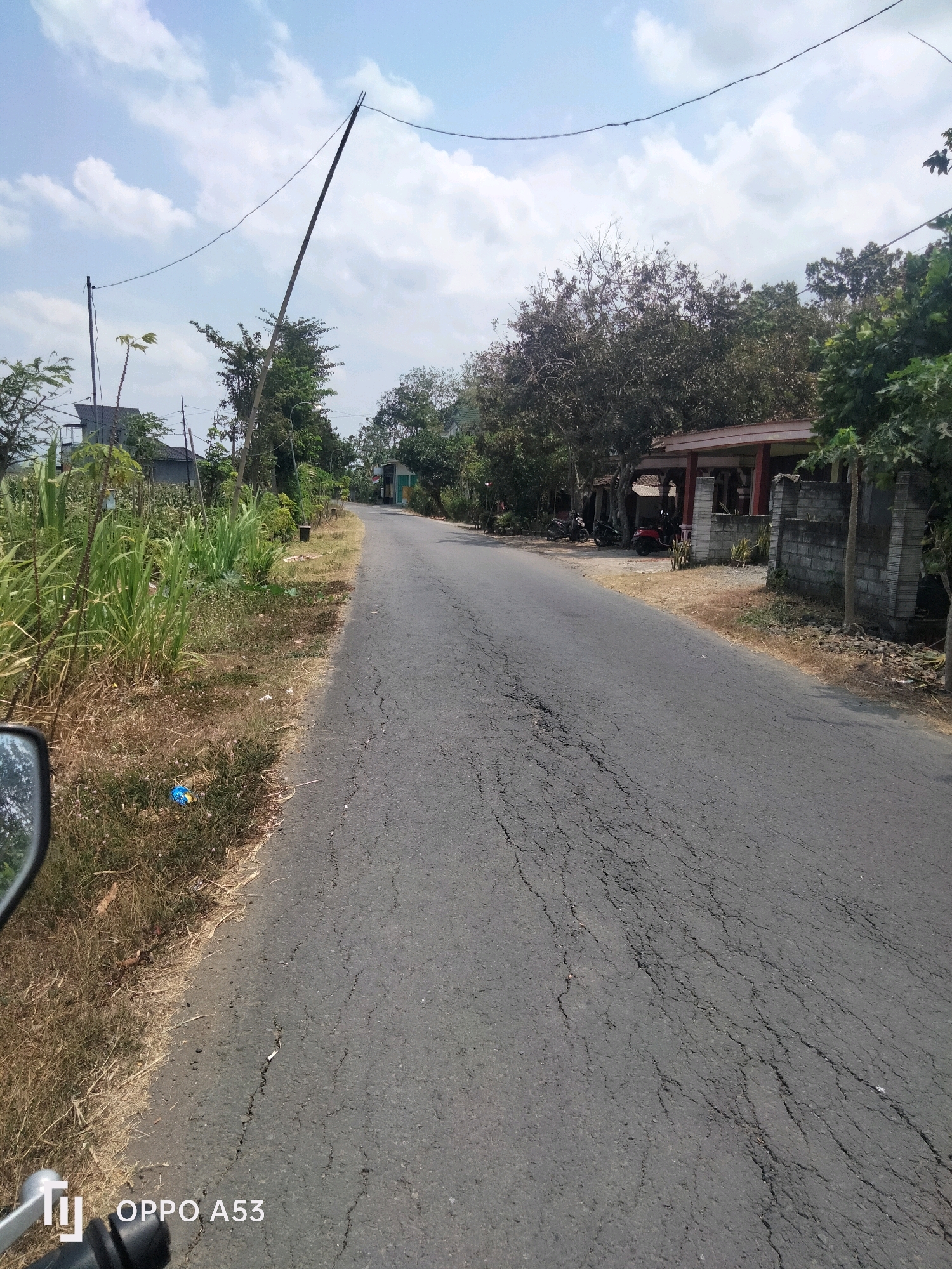 A paved road runs through a semi-rural area under a partly cloudy sky. The road has visible cracks and is lined with dry grass and some green vegetation on the left. On the right side of the road, there is a house with a red roof and pillars, bordered by a stone wall. Several motorcycles are parked in front of the house. Tall trees with dense foliage are scattered throughout the scene, and power lines stretch across the sky, supported by a tall, slender pole. The foreground shows part of a motorcycle's rearview mirror and a timestamp indicating "OPPO A53".