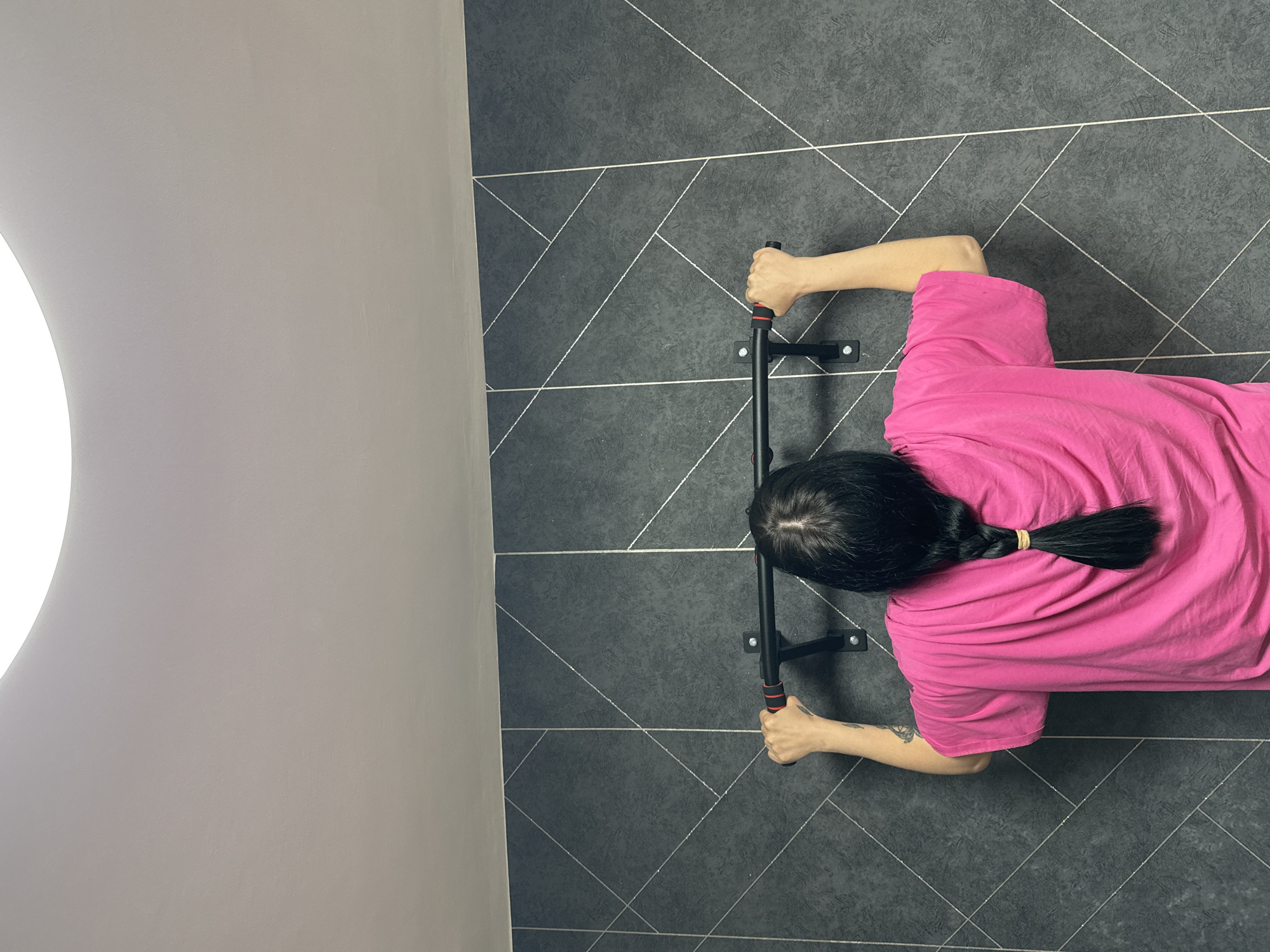 The image depicts a person, likely female based on the hair and clothing style, in a supine position holding onto a mounted pull-up bar. The person is wearing a pink t-shirt and has long, dark hair braided with a hair tie. The pull-up bar is black with red accents on the grips and is mounted on a dark gray wall with a geometric pattern. The adjacent wall is a light gray color, possibly indicating an indoor setting. The location is not discernable from the image.