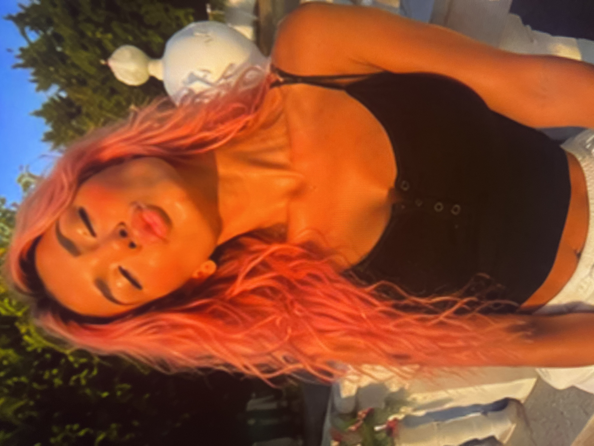 A woman with vibrant pink, wavy hair puckers her lips towards the camera, her eyes closed. She wears a black camisole with a four-button placket. The setting appears to be outdoors, possibly a garden or patio, with lush green trees and a clear blue sky in the background. A white decorative structure, possibly a pedestal or statue, is visible to her right. The lighting suggests it is late afternoon or early evening, casting a warm, golden glow on her skin and hair. The overall impression is one of a casual, posed portrait.