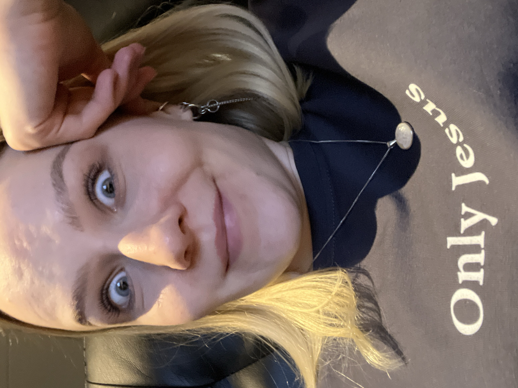 A woman with blonde hair and blue eyes is looking at the camera. She is wearing a gray shirt with the words "Only Jesus" printed in white. Her hand is raised near her face. She is also wearing a silver necklace. The photo appears to be taken indoors, with a leather or vinyl surface visible in the lower left corner. The image does not provide enough information to determine the location in Khimki, Russia.
