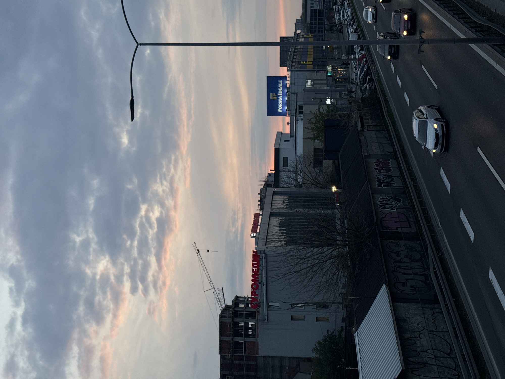 The image shows an urban landscape in the Vozdovac Urban Municipality, Serbia, at dusk. A six-lane highway runs parallel to buildings. Cars are driving on the highway. A construction site with a crane is visible on the left. Several buildings with signs, including one for "FORMA IDEALE" and another that appears to read "EURO MEDIK" are visible. The sky is cloudy with streaks of pink, indicating a sunset or sunrise. A street light pole is in the foreground. There is graffiti on the retaining wall.