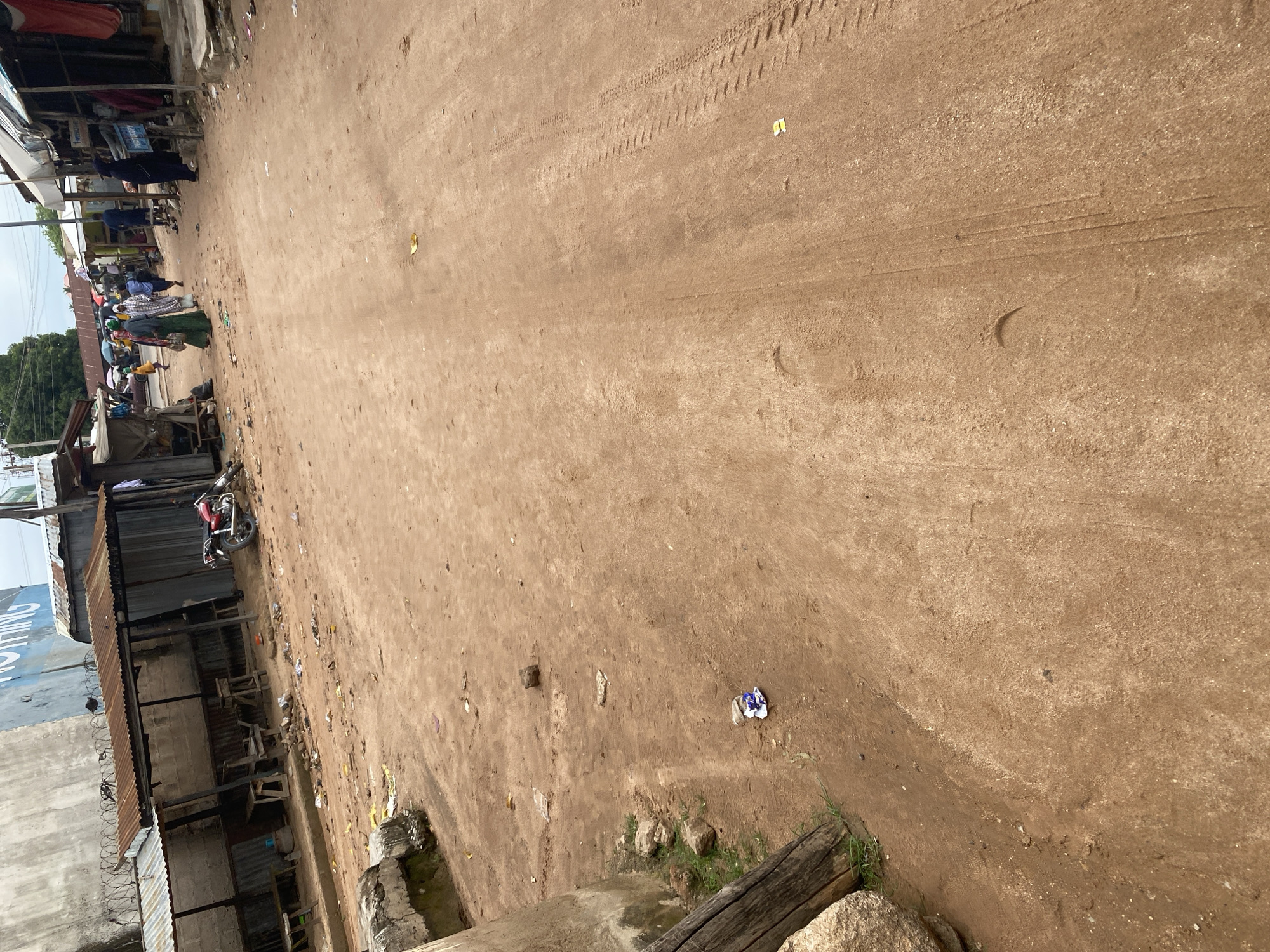 The image depicts a dusty, unpaved street in Jalingo, Nigeria, lined with informal market stalls and structures constructed from corrugated metal and concrete blocks. Several individuals are visible in the background, some walking and others gathered around stalls. A red motorcycle is parked near one of the structures. The ground is covered in reddish-brown dirt, with scattered debris and tire tracks from vehicles. The sky is overcast, suggesting a cloudy day. The overall scene conveys a sense of a bustling, everyday market environment in an urban setting in Nigeria.