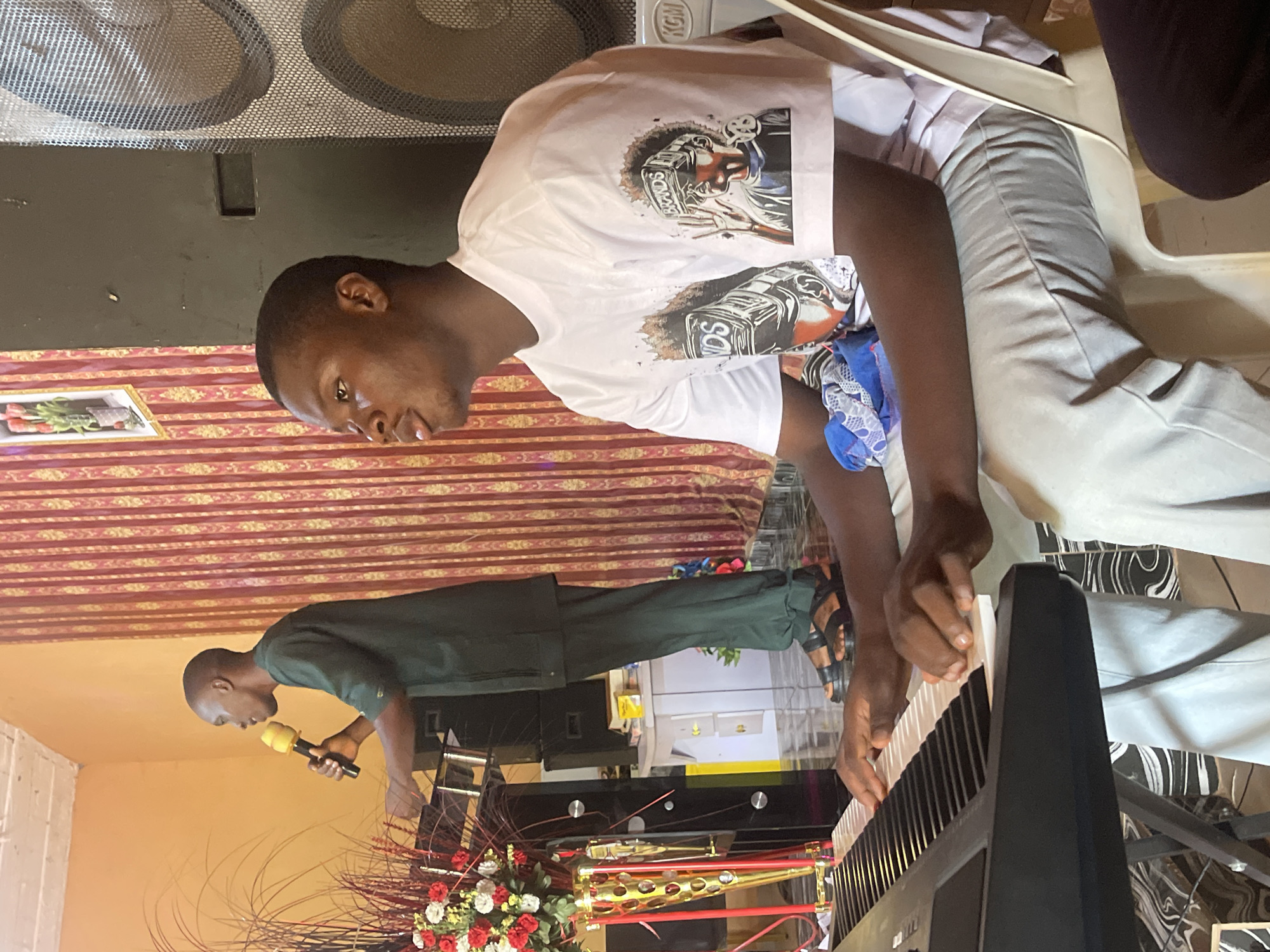 A medium, indoor shot depicts two people in a room where one person is playing a keyboard while another person sings into a microphone.
The person playing the keyboard is a dark-skinned man in a white, graphic t-shirt and light gray pants sitting on a white chair. His fingers are on the keyboard keys as he looks forward.
The singer is a dark-skinned man in a green shirt and pants holding a microphone. He stands near a flower arrangement on a stand.
The background includes speakers, patterned walls, and an object framed on the wall. The location is Unknown, Unknown.