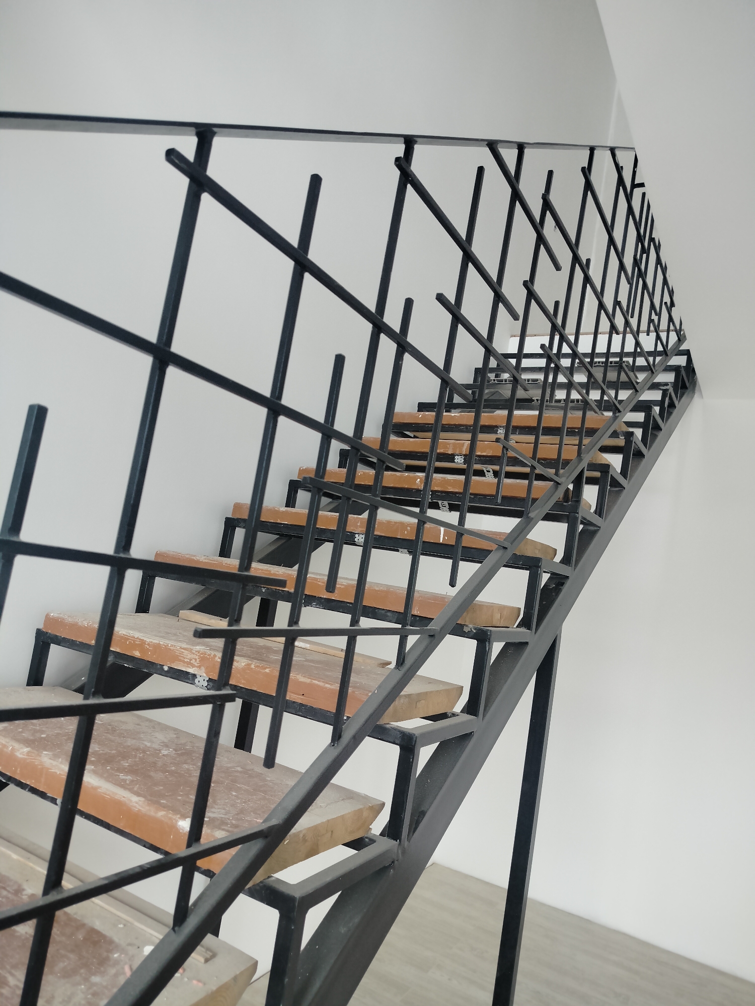 The image shows an indoor staircase with a metal railing in a building located in Прикамский, Russia. The staircase has wooden steps, and the railing is made of black metal bars in a lattice pattern. The walls are white. There are no people visible in the image. The lighting is bright, suggesting daytime. The floor is gray tile. The staircase appears to be newly constructed or recently renovated.