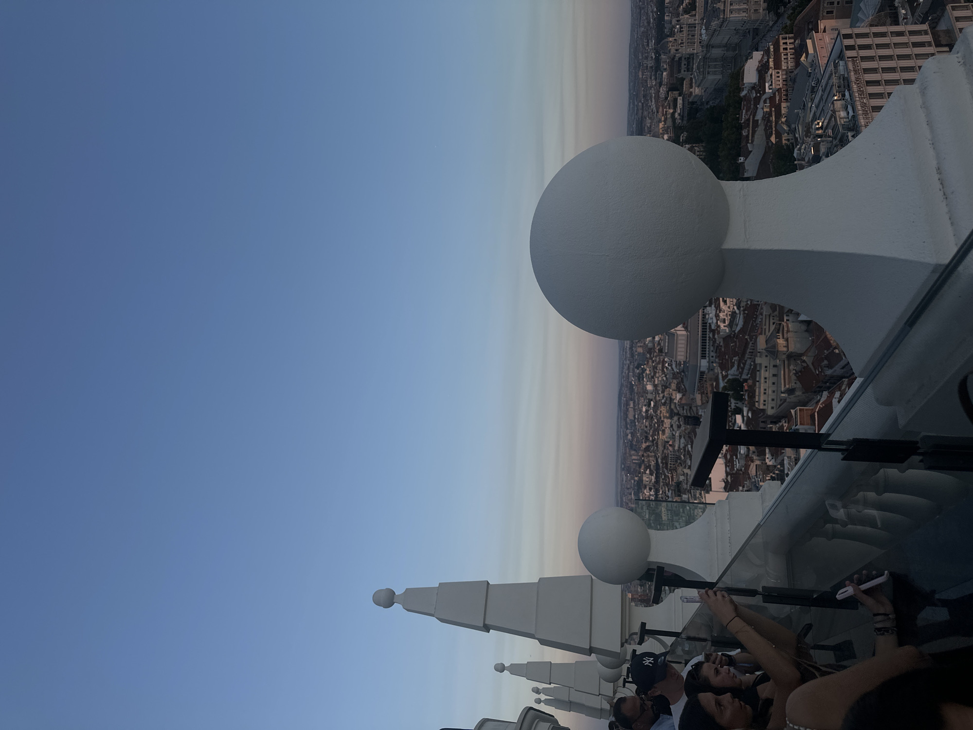 This is a photograph taken from a rooftop vantage point in Madrid, Spain, likely at dusk, given the soft light and gradient sky. In the foreground, a white decorative balustrade with spherical finials lines the edge of the rooftop. Several people are visible on the rooftop, some leaning over the balustrade, observing the city below. One woman in the lower left is holding a smartphone up, seemingly taking a picture or video. Another person is wearing a New York Yankees baseball cap. The cityscape of Madrid stretches out to the horizon, characterized by a dense collection of buildings with terracotta roofs and some larger, more modern structures. The sky transitions from a clear blue at the top to softer hues of pink and orange near the horizon, indicating the setting sun. The overall atmosphere suggests a social gathering or a tourist attraction offering panoramic views of the city.