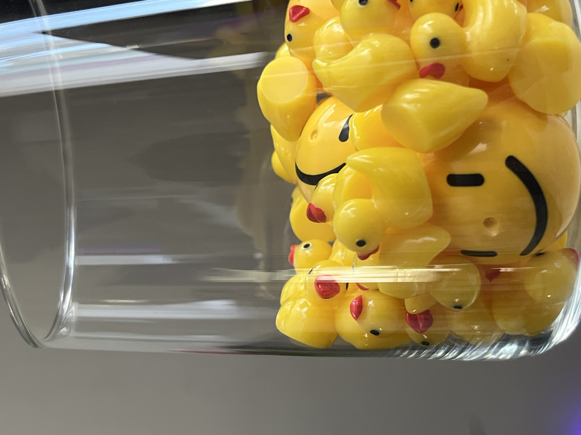 A clear glass vase is filled with numerous yellow rubber ducks. Some of the ducks have red beaks and black eyes. One duck in the foreground has a smiley face printed on it. The background is a plain, neutral color. The image does not contain information about the city of Babruysk, Belarus.