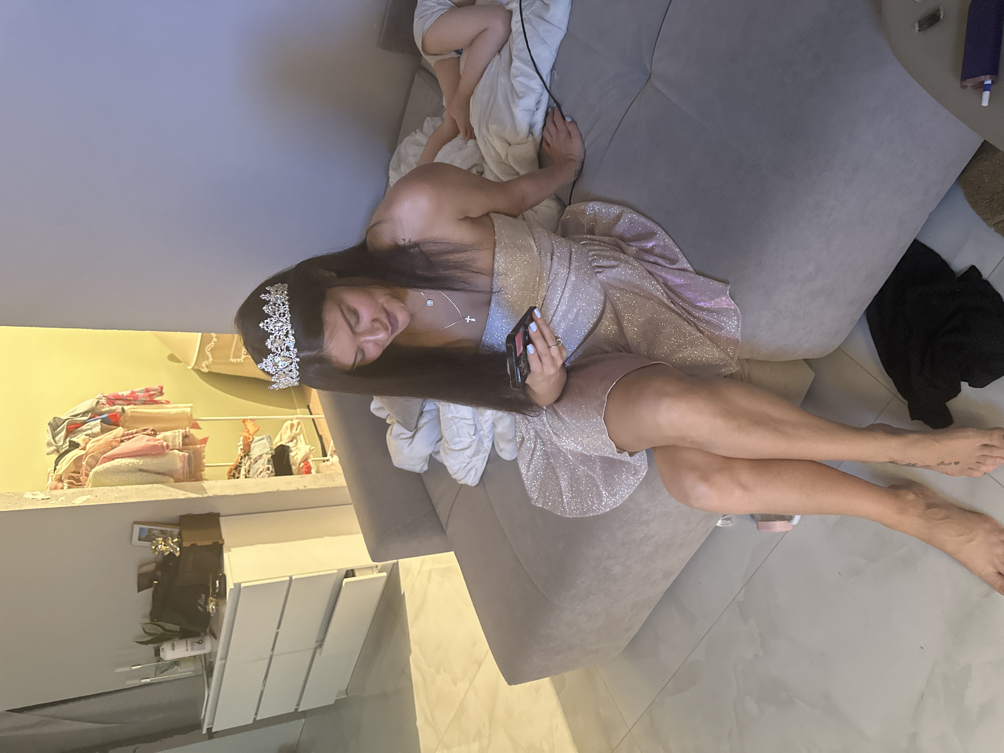 An adult woman is depicted reclining on a light-colored sofa in an indoor setting, likely a living room or bedroom in Moscow, Russia. She has long, dark hair and is wearing a sparkly, strapless dress, a tiara, and a necklace with a cross pendant. She holds a smartphone in her hands and looks down at it with a gentle smile. Her legs are bare, extending onto a light-colored tiled floor.
At the top left of the sofa, another individual, possibly a child, is partially visible, covered by a white blanket, with their legs showing. A charging cable runs across the blanket near them.
In the background, to the woman's left, a shelf or ledge holds neatly folded clothes or towels, alongside various personal items such as bottles and a small dark bag. Below this, a white dresser or cabinet with multiple panels is visible. On the floor to the woman's right, a black piece of fabric or clothing is present. The artificial lighting suggests the scene takes place in the evening or at night. The woman's festive attire, including the tiara, indicates a celebratory occasion. No discernible text is visible.