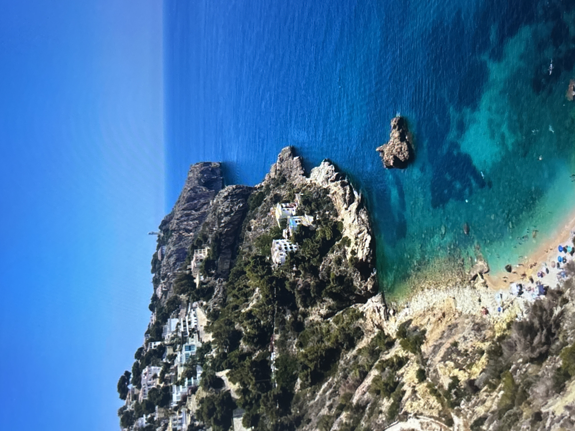 The image presents an aerial view of a picturesque coastal landscape, rotated 90 degrees counter-clockwise from a typical upright orientation. The left side of the frame is dominated by a clear, bright blue sky. A rugged landmass, characterized by steep cliffs and hills covered in lush green vegetation, occupies the central and lower portions of the image.
Numerous white and light-colored buildings, appearing to be houses or villas, are strategically nestled into the hillsides and perched on the clifftops. Some of these structures feature visible swimming pools. A path or road winds through the densely vegetated terrain where the buildings are situated.
To the right, a vast expanse of ocean transitions from deep blue in the distance to a vibrant turquoise closer to the shore, indicating shallow, clear waters that reveal the rocky seafloor. Several dark, prominent rock formations protrude from the water. In the bottom right corner, a small, crescent-shaped beach made of light-colored pebbles or sand is visible. Small figures, identified as people, are scattered on the beach, some near what appear to be colorful umbrellas or towels, suggesting sunbathing or other leisure activities.
The scene is illuminated by bright daylight, with clear skies and strong sunlight, indicating midday or early afternoon. The overall impression is that of an idyllic Mediterranean-style coastal destination, characterized by natural beauty and human settlements integrated into the landscape. No visible text is present in the image.