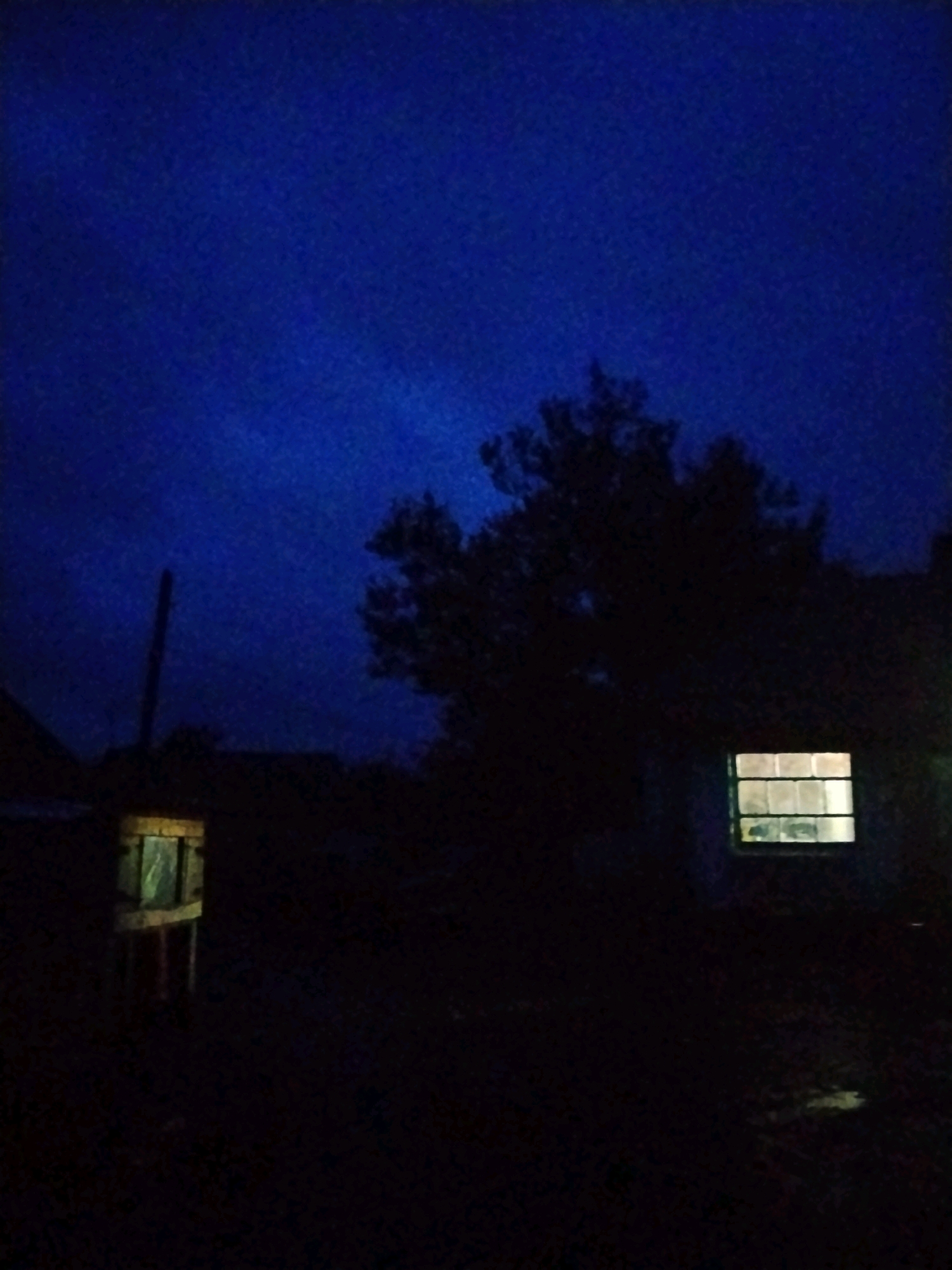 In the Ustyvytsia, Ukraine, a dimly lit outdoor scene unfolds under a deep blue twilight sky. In the foreground, a weathered wooden structure with a glass pane stands on the left. To the right, a tree with dark foliage is silhouetted against the sky, and a window with illuminated panes glows from within a structure, suggesting occupancy. The ground is dark and indistinct.