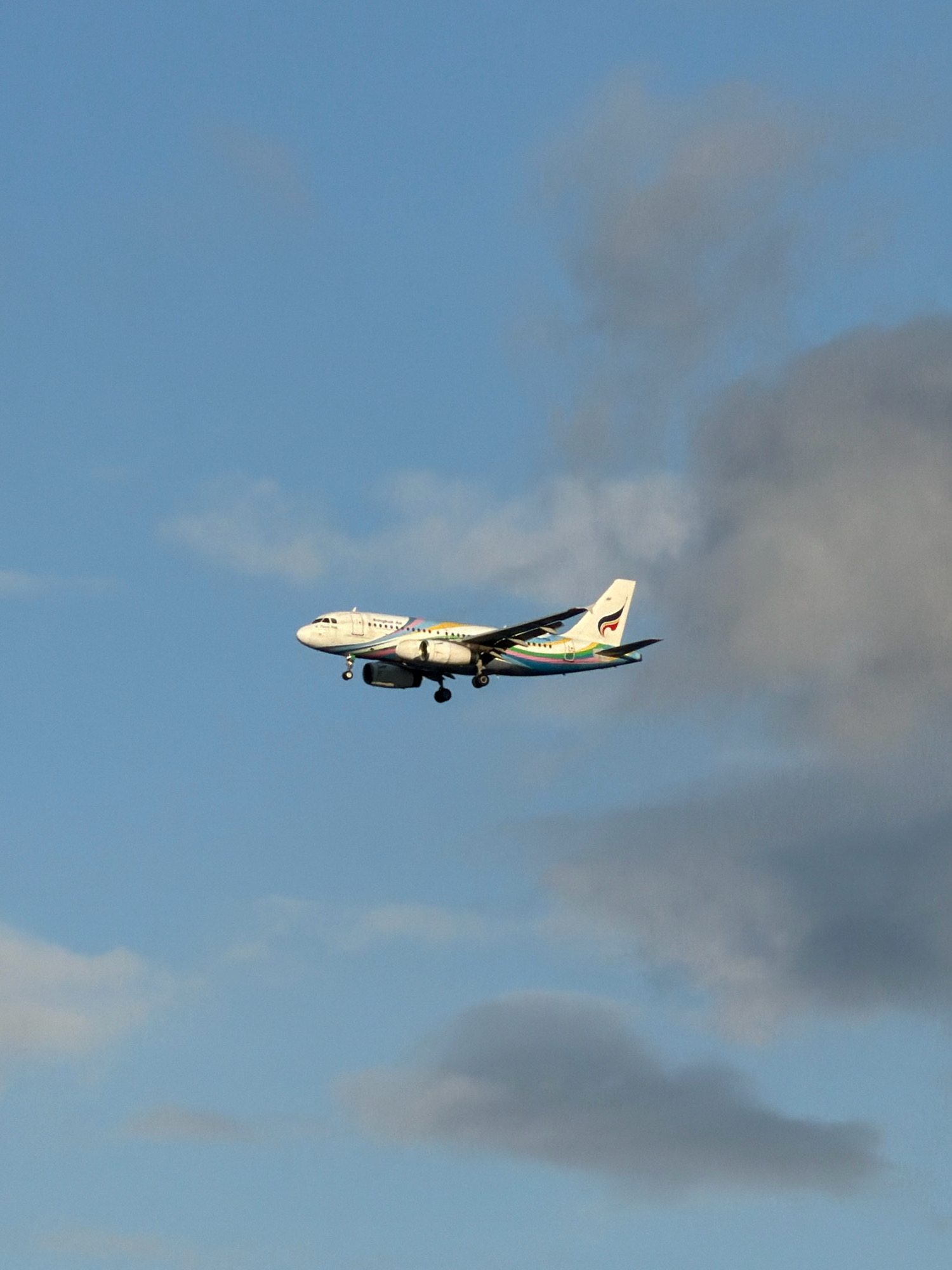 A white passenger airplane, identified by the "Bangkok Air" logo on its tail fin, is captured mid-flight against a sky of blue with scattered gray clouds. The airplane appears to be on a descent, as its landing gear is deployed and its engines are visible beneath the wings. The fuselage is decorated with a colorful stripe that transitions through shades of blue, green, pink, and yellow. The image was taken from a low angle, looking up at the aircraft. The lighting suggests it is daytime, possibly late afternoon or early morning, given the soft light. The location is specified as the sky above Ko Samui District, Thailand.