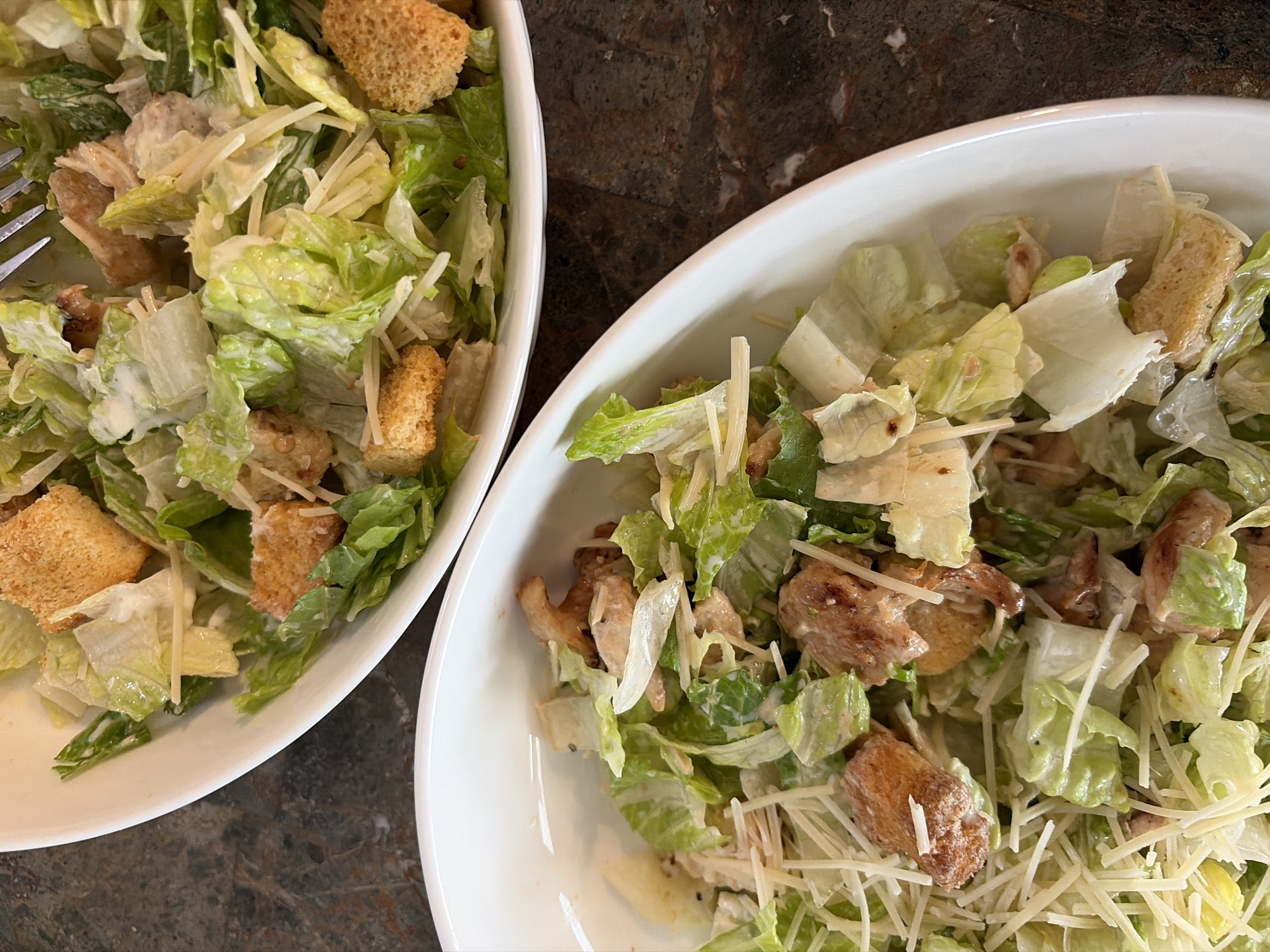 Two white bowls containing Caesar salad are positioned on a dark brown marbled surface. The salad consists of chopped romaine lettuce, croutons, parmesan cheese shavings, and Caesar dressing. A fork is visible in the bowl on the left. The scene is likely indoors, given the controlled lighting and table setting. The location details "Trophy Club, United States" suggest a domestic or restaurant setting within that area.