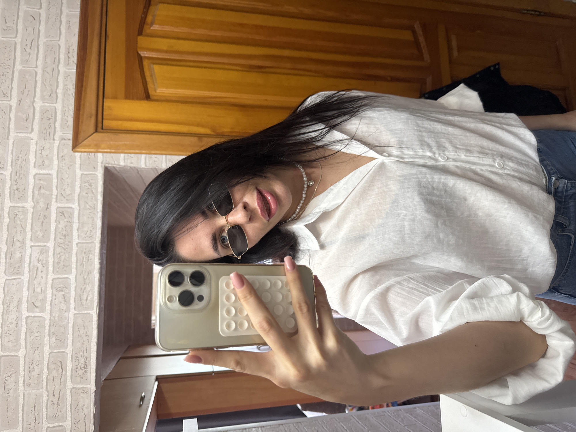 A young woman with long, dark hair and blue eyes is taking a selfie in a room. She is wearing a white, oversized button-down shirt, blue jeans, sunglasses with gold frames, and two necklaces: one with pearls and a small pendant, and another thin gold chain. She is holding a smartphone in her right hand. The background shows a wall with white brick wallpaper, a wooden door, and a reflection in a mirror. The scene appears to be an indoor residential setting, potentially an apartment, located in the city of Chaikovsky, Russia.