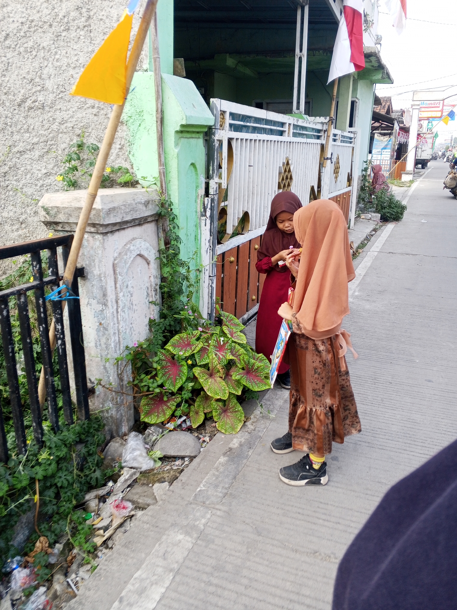 Two young girls, wearing hijabs and traditional Indonesian clothing, stand on a sidewalk in front of a building. The girl on the left wears a maroon dress, and the girl on the right wears a brown patterned dress. The girl on the left is holding something small in her hands, and the girl on the right is holding a book with a colorful cover. In the background, a white fence with ornate details separates the sidewalk from the building, which appears to be a shop or residence with a green facade. Indonesian flags are visible on the building's upper level. The street is paved with asphalt, and there are some plants and debris along the edge of the sidewalk. A yellow flag on a wooden pole stands to the left of the girls. The scene is consistent with a residential or commercial street in Ciruas, Indonesia.
