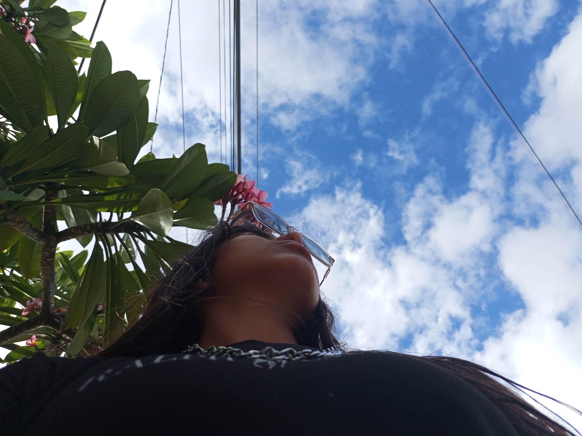A low-angle shot captures a person wearing sunglasses and a chain necklace, positioned beneath the foliage of a tree with pink flowers, against a backdrop of a blue sky dotted with white clouds. Power lines are visible stretching across the sky. The location is presumed to be Juan Griego, Venezuela.