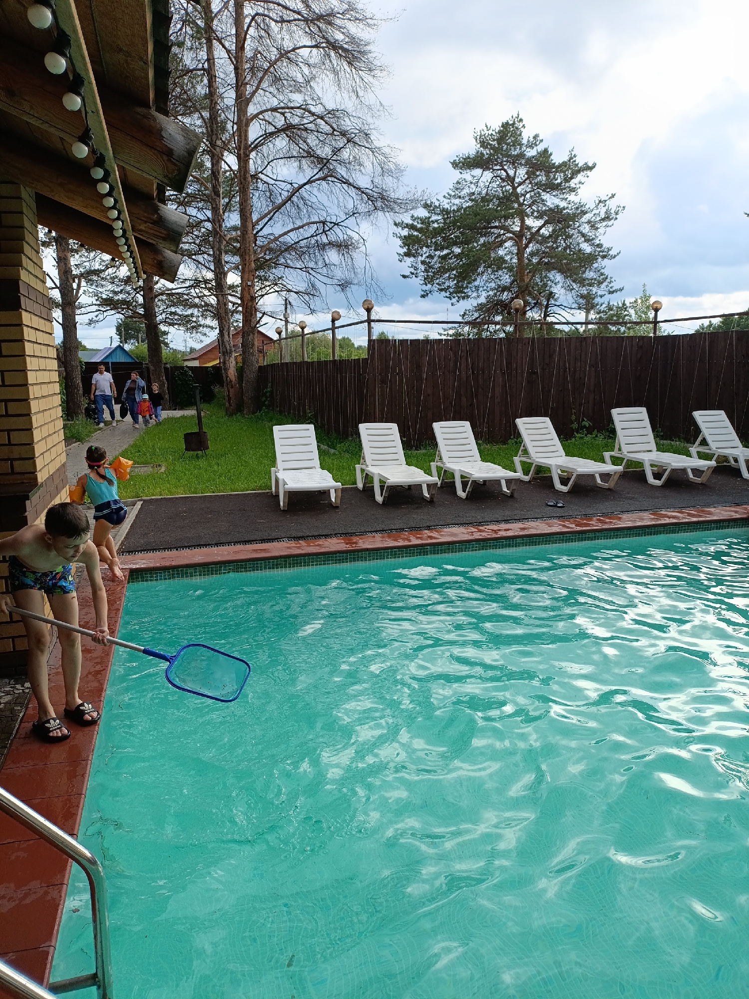 This image captures an outdoor swimming pool area in Chaikovsky, Russia, under an overcast sky. In the foreground, a young boy in patterned swim trunks and black Adidas slides stands at the edge of the turquoise pool, holding a blue net attached to a long pole, reaching into the water. Behind him, another child in a blue swimsuit and orange arm floats walks away from the pool.
The swimming pool occupies the mid-ground, featuring clear, rippling water. Along the opposite side of the pool, five white plastic sun loungers are neatly arranged on a dark, paved surface. A brown wooden fence, topped with sphere-shaped lights, runs along the back of the pool area, separating it from a more wooded landscape.
To the left, a portion of a brick building with wooden eaves adorned with a string of globe lights is visible. A paved pathway extends from this structure into the background, where a group of people, including several adults and a small child, are seen walking. The background is characterized by numerous tall trees, some with bare branches and others with green foliage, against a cloudy, bright sky. A small blue building and another wooden structure are faintly visible in the distance among the trees.
The scene suggests a relaxed daytime recreational activity, possibly late afternoon given the soft lighting. The children appear to be enjoying the pool area, with one actively engaged in skimming the water. The overall atmosphere is tranquil and inviting for outdoor leisure.