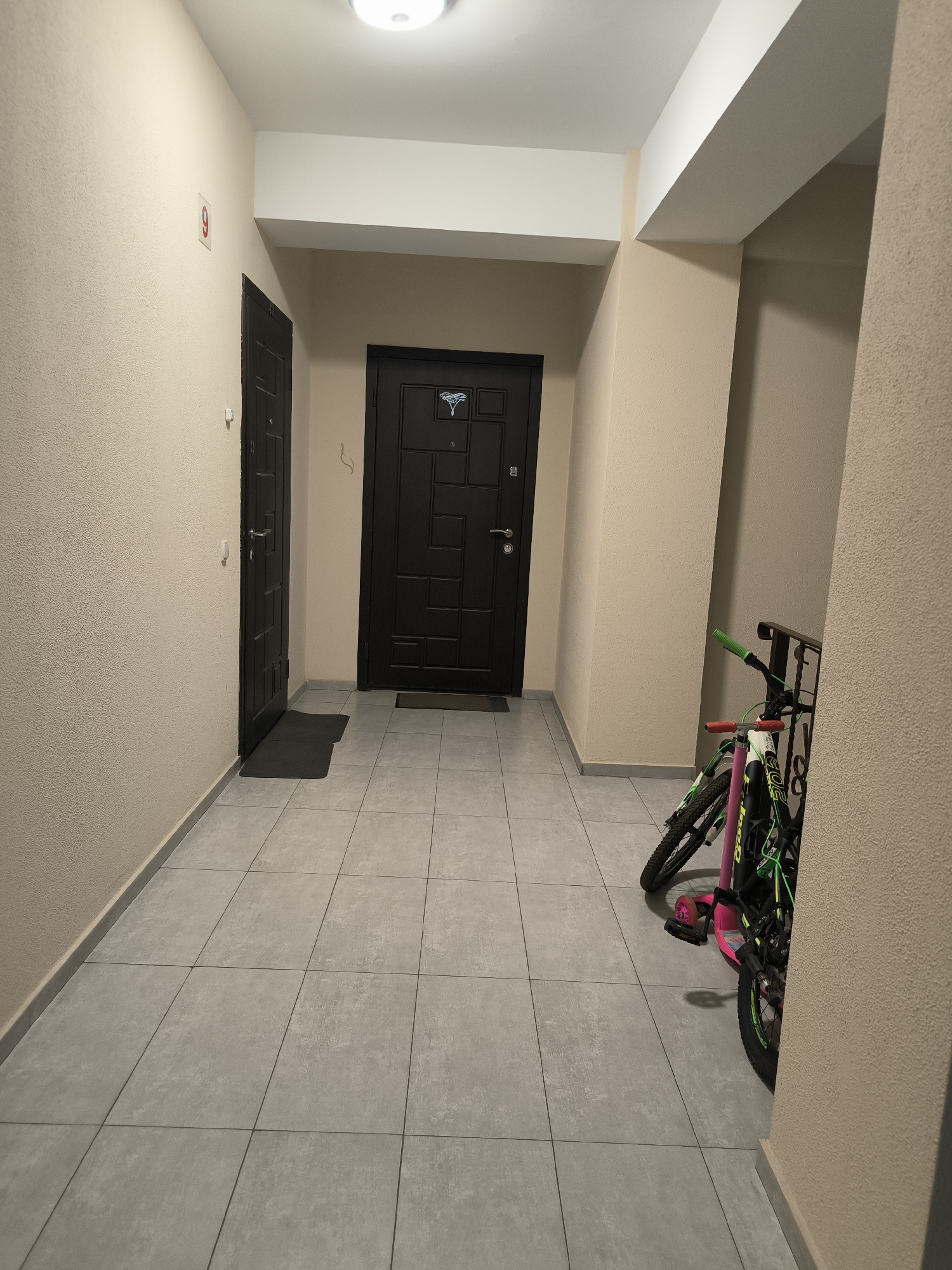 The image shows a dimly lit indoor hallway in an apartment building. The hallway has tiled floors and beige textured walls. There are two dark-colored doors visible. On the left wall, a red sign with the number "9" is displayed. On the right side of the hallway, two bicycles are parked, one primarily pink and white with green accents, and another partially obscured by the first, also with green accents. The environment appears to be a common area within a residential building, likely in Chișinău, Moldova, given the context. No people are present in the image.