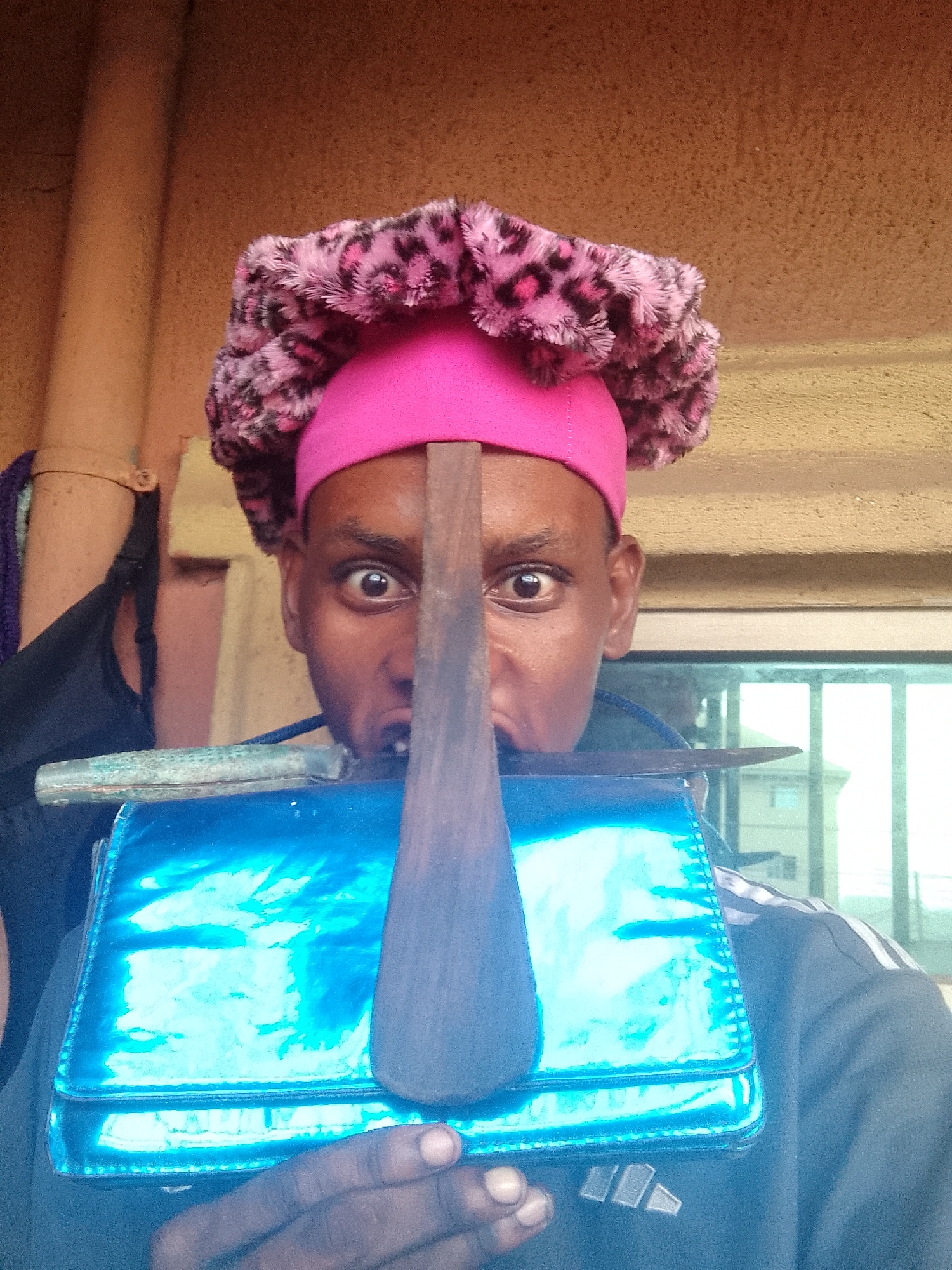 A person is holding a shiny blue purse and a knife in front of their face. The person is wearing a pink hat with a furry leopard-print trim. The person's eyes are wide and they appear to be looking directly at the camera. The background is a wall with a pipe running vertically. There is also a window in the background.