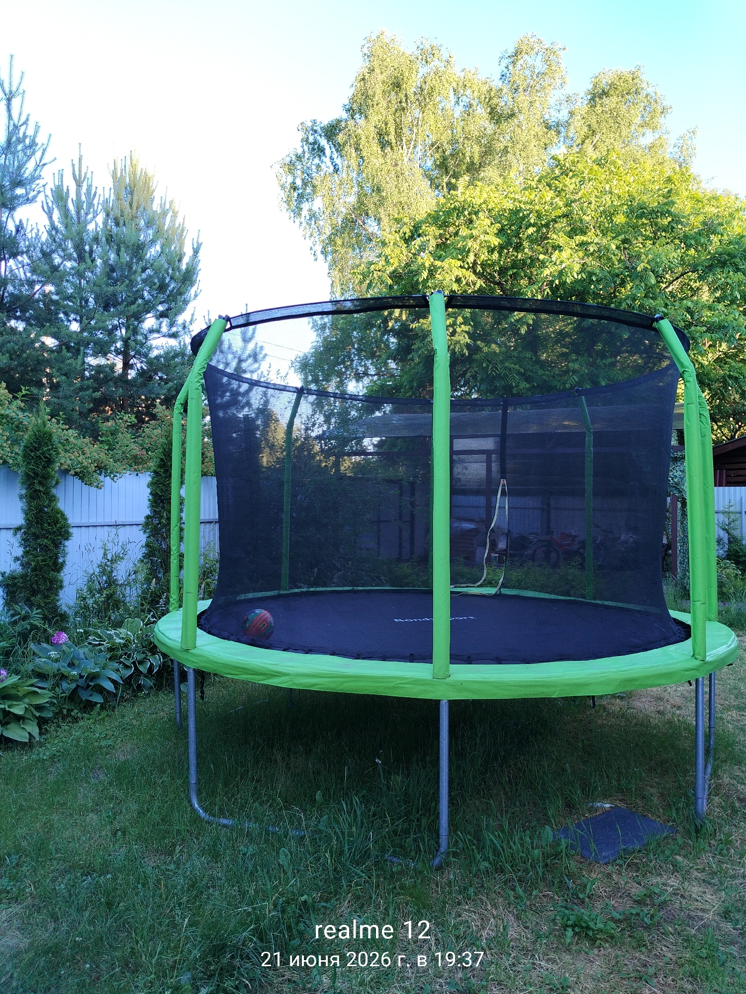 A trampoline with a green frame and a black safety net is set up in a grassy backyard. A multi-colored ball rests on the trampoline mat. In the background, there are trees, a fence, and some buildings. The sky is clear and blue. The image also contains a watermark indicating "realme 12" and the date "21 июня 2026 г. в 19:37". The location is identified as Космос-2 Снт, Russia.