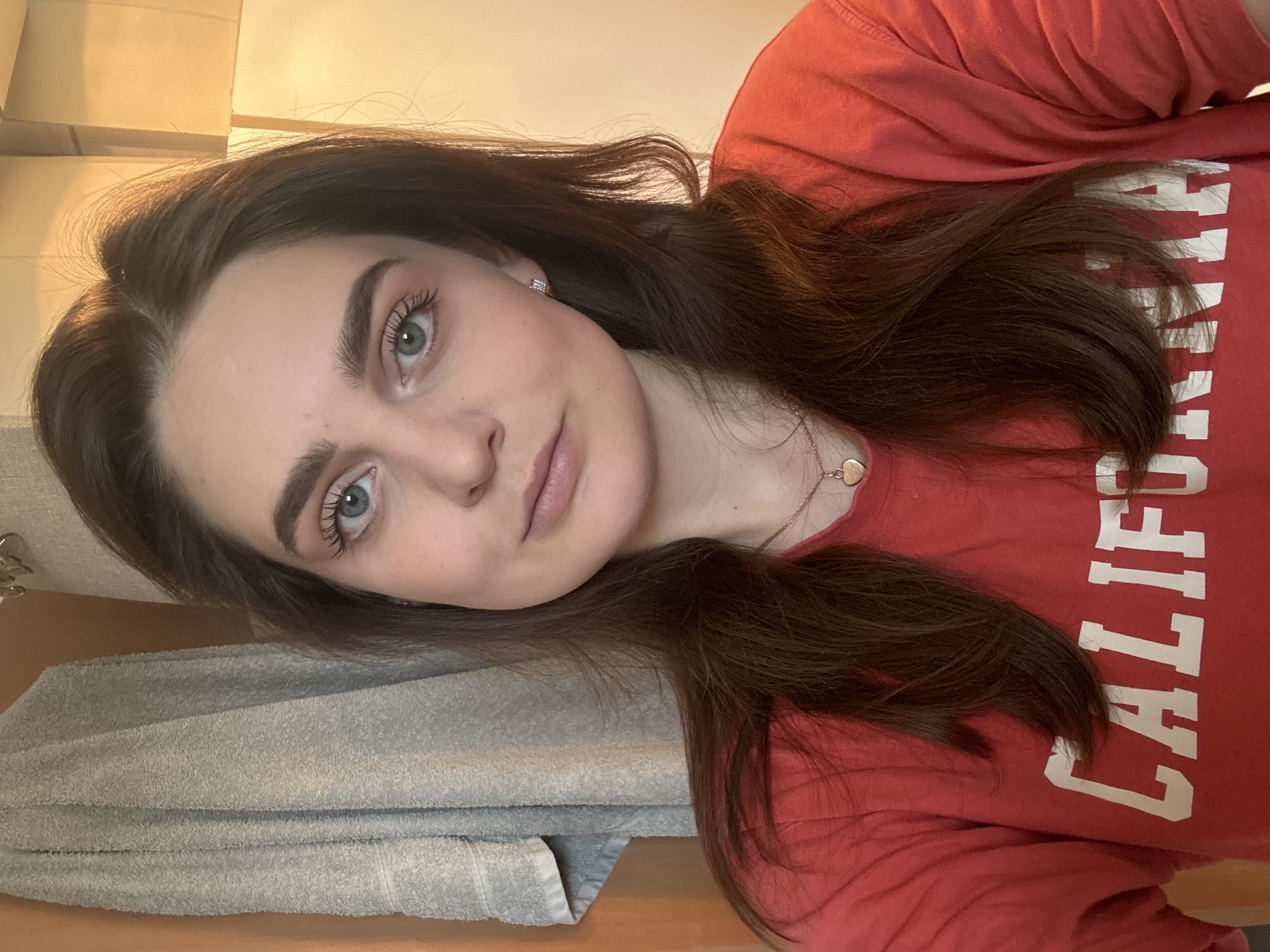 An indoor, close-up portrait features a young adult female, with the image oriented sideways. She has long, dark brown hair, parted on her left, falling across her shoulders and chest. Her light blue-grey eyes gaze directly at the camera, conveying a neutral to slightly serene expression with a subtle hint of a smile. Her eyebrows are well-defined, and her eyelashes show visible mascara. She is wearing small, square stud earrings and a delicate gold-colored necklace with a heart pendant.
She is dressed in a red long-sleeved shirt, which features white capital letters. The visible text reads "CALIFORNI", suggesting the word "CALIFORNIA". In the background, a neatly folded grey towel is visible on a surface, likely a counter or piece of furniture. The upper background consists of a plain, light-colored wall or surface, with warmer, golden-beige tones indicating artificial indoor lighting. The scene appears to be a casual setting, possibly a private residence in Hainburg an der Donau, Austria. The image captures a posed moment, focusing on the individual's facial features and upper body. The time of day and external weather conditions are not discernible.
