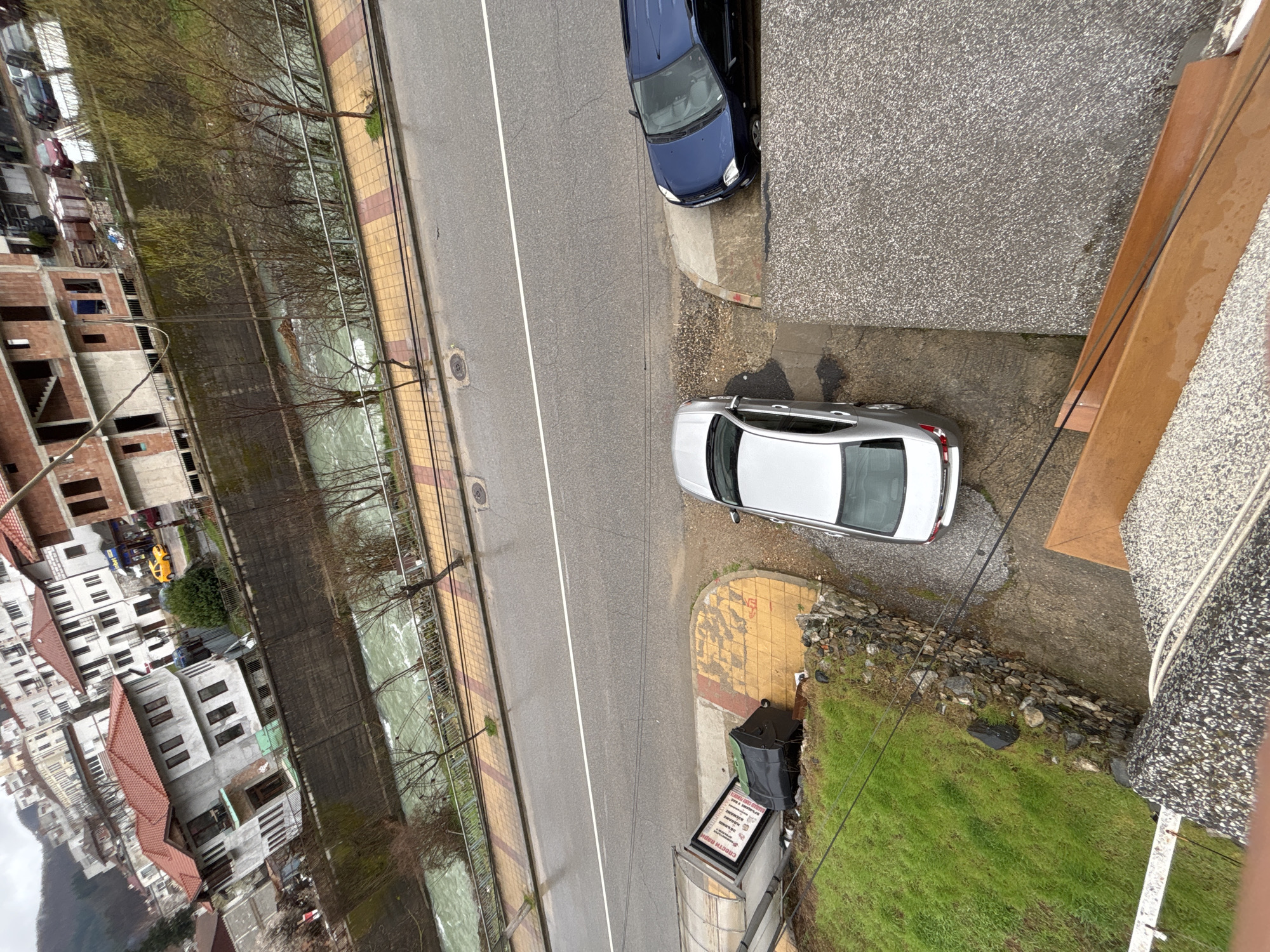 The image is a high-angle view of a street scene in Zlatograd, Bulgaria. A two-lane road runs parallel to a river. Buildings, including an unfinished construction site, are visible on the opposite side of the river. On the right side of the road, there is a silver car parked on the sidewalk next to a trash can and an advertising sign. A blue car is parked further up the road, also on the sidewalk. The terrain on the right side of the road slopes downwards, covered with grass and some stone walls. A portion of a building or balcony is visible in the lower right corner of the image. The weather appears overcast.