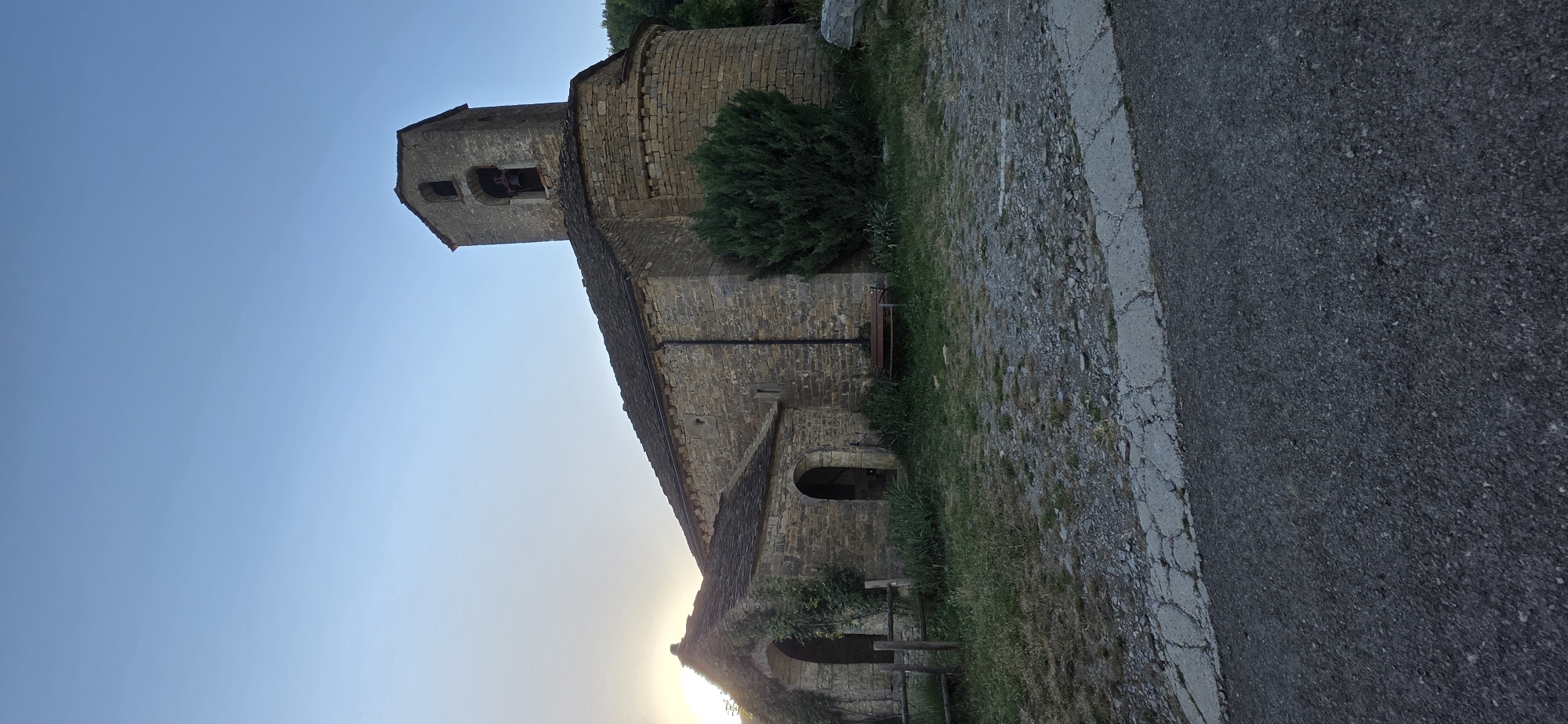 An exterior, low-angle, wide shot captures a stone church with a bell tower against a clear blue sky at dusk. The church, constructed from rough-hewn stones, features a prominent bell tower with two open belfry arches and a bell inside. A rounded apse is visible on the right side of the building, and a smaller, attached structure with arched doorways stands to the left. The building's roof is tiled with what appears to be slate.
In front of the church, a patch of dry grass and gravel surrounds the structure. A wooden fence is positioned to the left of the entrance, and a dark wooden bench sits in the grass near the right side of the building. A tall, dense evergreen tree is situated to the right of the main church structure. The ground in the foreground is a mixture of gravel and cracked concrete, transitioning into a darker asphalt road. The sun is low on the horizon, casting a soft light on the scene, particularly illuminating the left side of the church where ivy grows over an archway.
The location is the Church of San Cipriano in the village of Gavín, a municipality within Biescas, Spain.