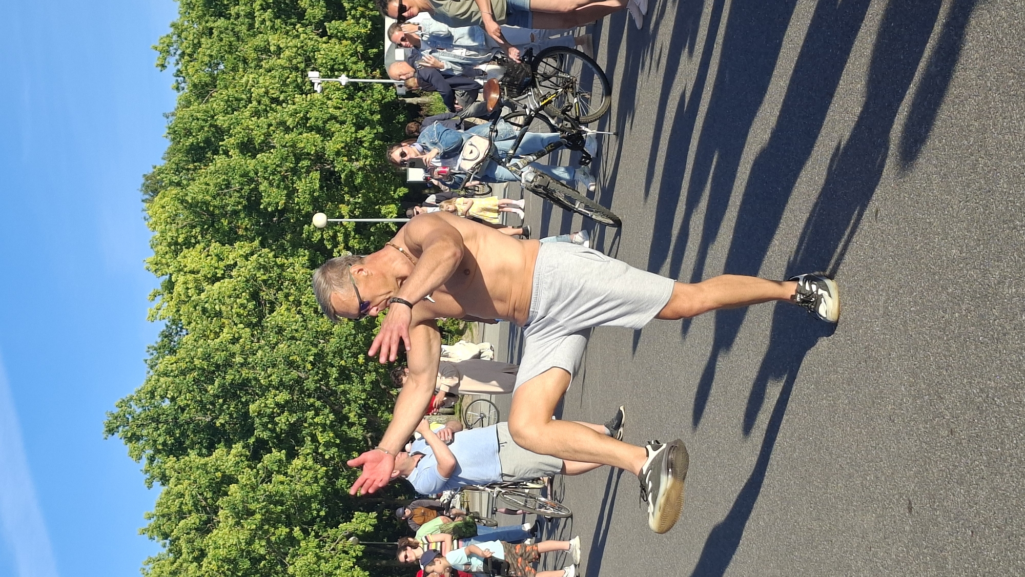 A shirtless man in grey shorts and athletic shoes performs a martial arts pose with his right leg extended forward and his arms outstretched in a public outdoor space. He is wearing sunglasses and a necklace. Behind him, a crowd of people are visible, some holding phones and cameras, suggesting an event or performance. Several bicycles are parked in the background. The environment appears to be a park or promenade, with a line of green trees and a clear blue sky overhead. The scene is lit by bright sunlight, casting long shadows on the asphalt ground. The location is in Tallinn, Estonia, specifically within the area of the Tallinn Song Festival Grounds, based on the architecture and vegetation visible in the image.