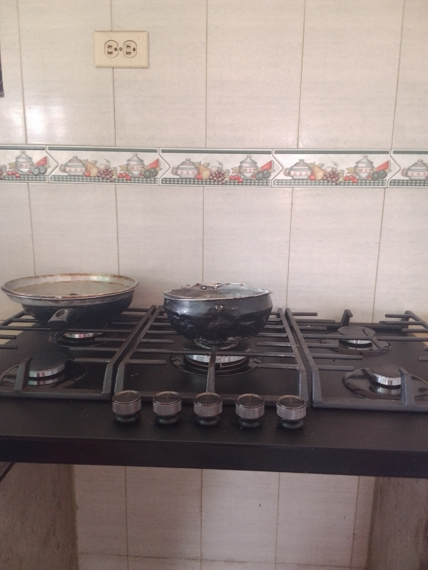 The media file captures a close-up view of a modern gas stovetop in what appears to be a kitchen setting. The stovetop is black with a metal grate on top, and there are four silver knobs on the front panel. Two burners are occupied: a large, dark pot with a lid sits on the right burner, and a wide, shallow pan is on the left burner. The pan contains some liquid, suggesting it's in use or has recently been used. The background features a tiled wall with a decorative border of fruit and pottery motifs. Above the border, a light-colored electrical outlet with two sockets is visible, mounted on a tiled section of the wall. The lighting suggests daytime. The overall impression is of a functional kitchen space, possibly in a home or a small eatery in La Tagua, Venezuela. No people are visible, and there is no visible text in the image.